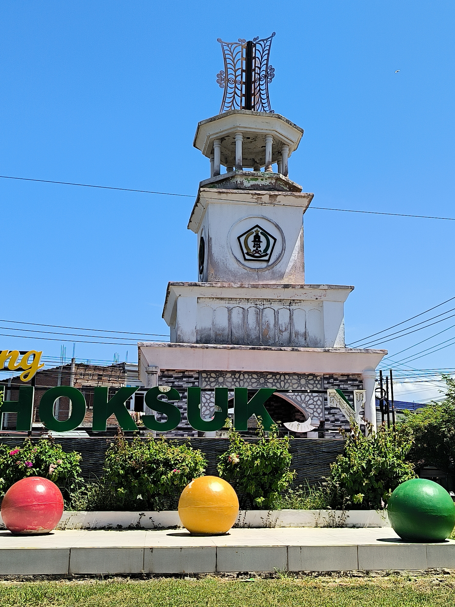 The image shows a sign with the name "Dayahlhoksukon" in green letters, with a tower structure behind it. The tower is white and has a small, ornate metal structure on top. There is a round emblem on the tower with green and black details. In front of the sign are three large, painted spheres: red, yellow, and green, placed along a concrete border with greenery in between. The sky is clear and blue. Overhead power lines are visible above the tower and surrounding buildings. The location is Dayahlhoksukon Barat, Indonesia.