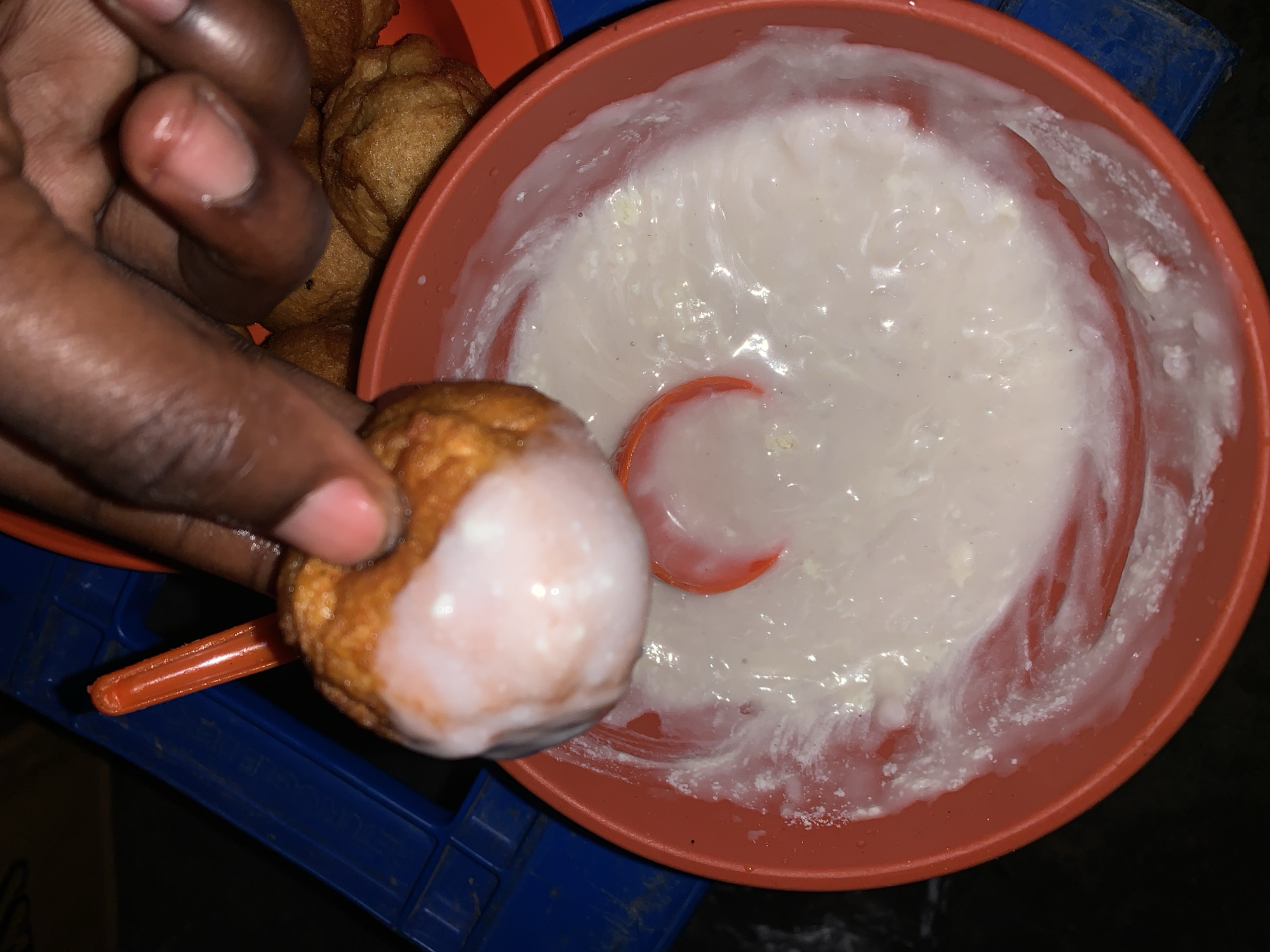 A close-up, high-angle shot captures a person's dark-skinned hand dipping a golden-brown, fried dough ball into a bowl of white, creamy substance. The hand holds one dough ball, which is partially coated in the thick white mixture, while several other similar dough balls are visible in an orange container to the upper left. The main orange bowl contains the white liquid, which has a slightly textured, lumpy appearance, and a small orange spoon or scoop is submerged in it. The entire setup rests on a blue plastic crate or object, part of which shows faint, illegible text that appears to begin with "ENUKA" or similar letters. The lighting is somewhat dim, suggesting an indoor setting or evening time. The food items strongly resemble Nigerian street food, specifically puff-puff (the fried dough) often served with pap or akamu (the white, creamy substance), consistent with the given location context of New Stadium, Nigeria.