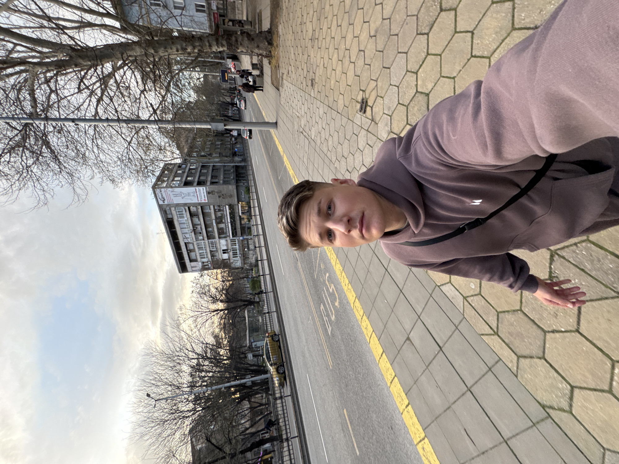 The image depicts a person standing on a sidewalk next to a street in Plovdiv, Bulgaria. The person is wearing a purple hooded sweatshirt with a black strap across their chest. The sidewalk is paved with hexagonal concrete tiles and is bordered by a yellow painted line marking the edge of the roadway. The street shows markings for a bus lane.
In the background, there is a multistory building with a large advertisement on its facade. Bare-branched trees line the street. Other cars and people are visible in the distance. The sky is overcast.
