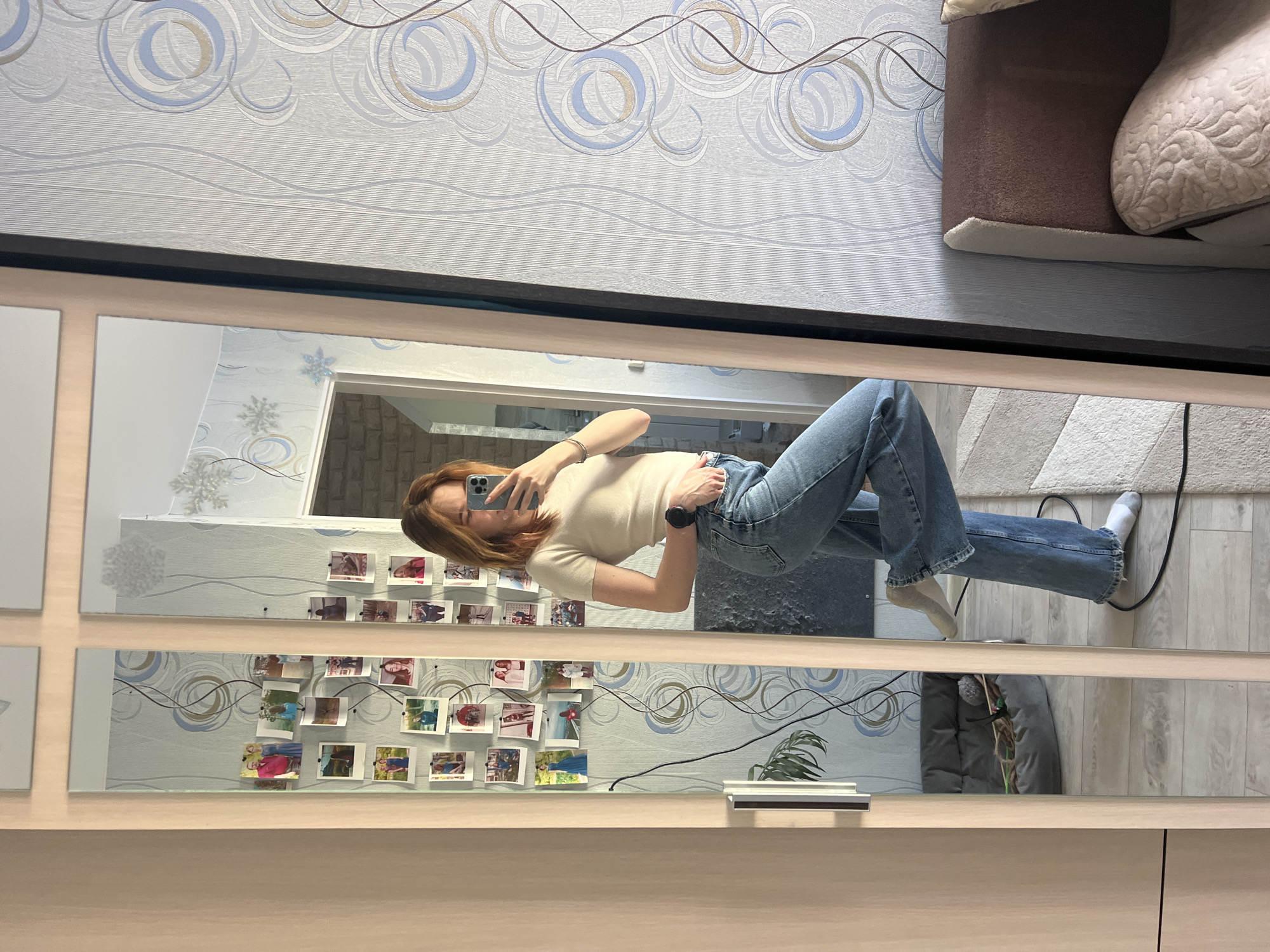 The image shows a reflection in a mirrored wardrobe, likely in a residential interior. A woman is holding a smartphone to take a picture of herself. She is wearing jeans and a light-colored t-shirt. The background visible in the mirror includes a wall with patterned wallpaper, a photo display board, and other domestic items, like a couch. The floor is light-colored wood. The picture was taken in an apartment in Chaikovsky, Russia.
