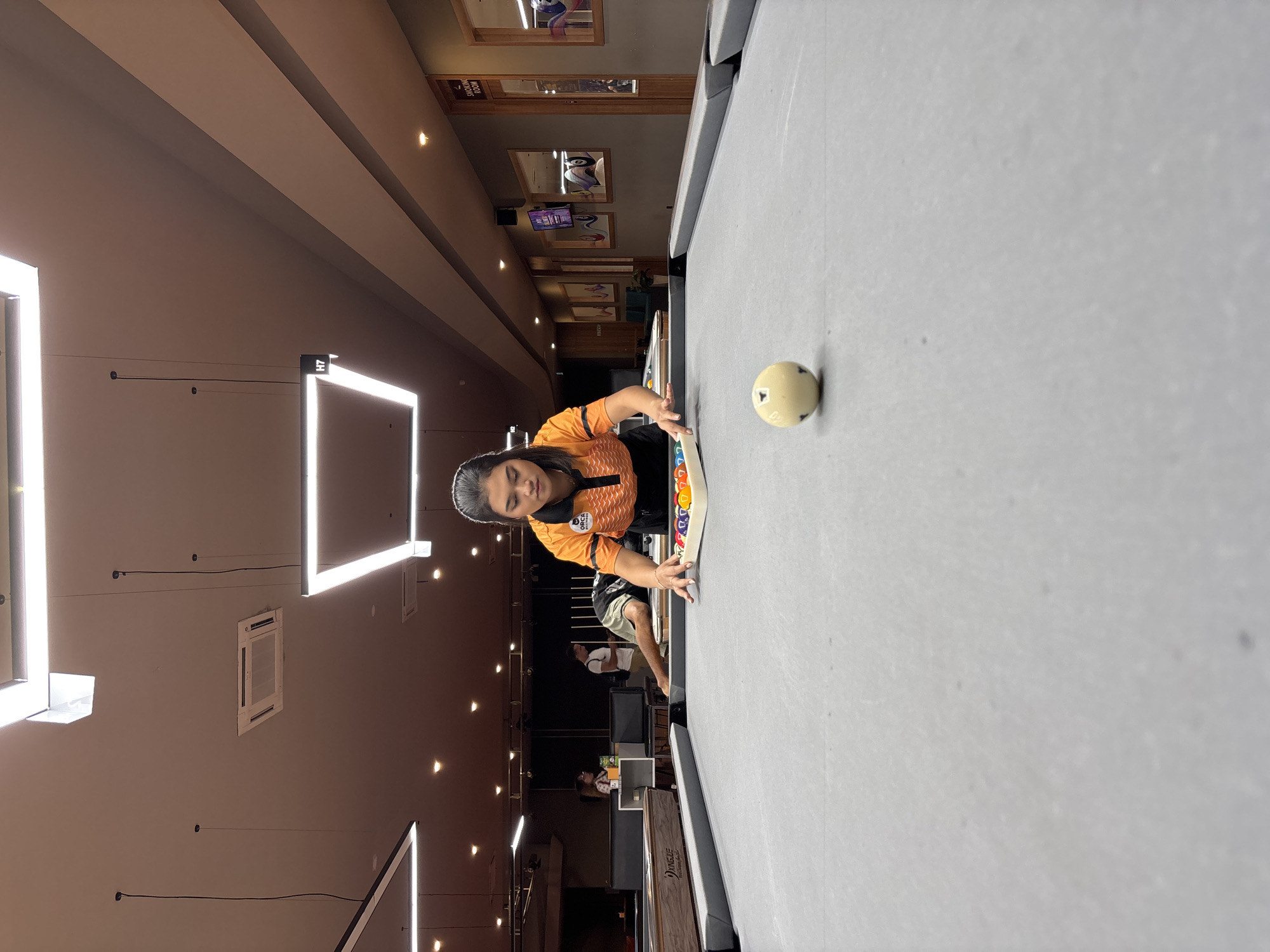 A woman is setting up billiard balls on a pool table. She is wearing an orange and black polo shirt with a logo on the left chest that says "ORCA BILLIARDS". She is in the process of arranging the balls in a triangular rack on the table. In the foreground, a cue ball is positioned on the felt of the pool table. The background of the image shows other pool tables, people playing, and wall decor. The environment appears to be an indoor billiard hall, likely in Kuta Selatan, Indonesia, given the presence of "SMOKING ROOM" signage on the wall, which is common in public establishments in Indonesia. The lighting is bright and consistent with indoor recreational facilities.