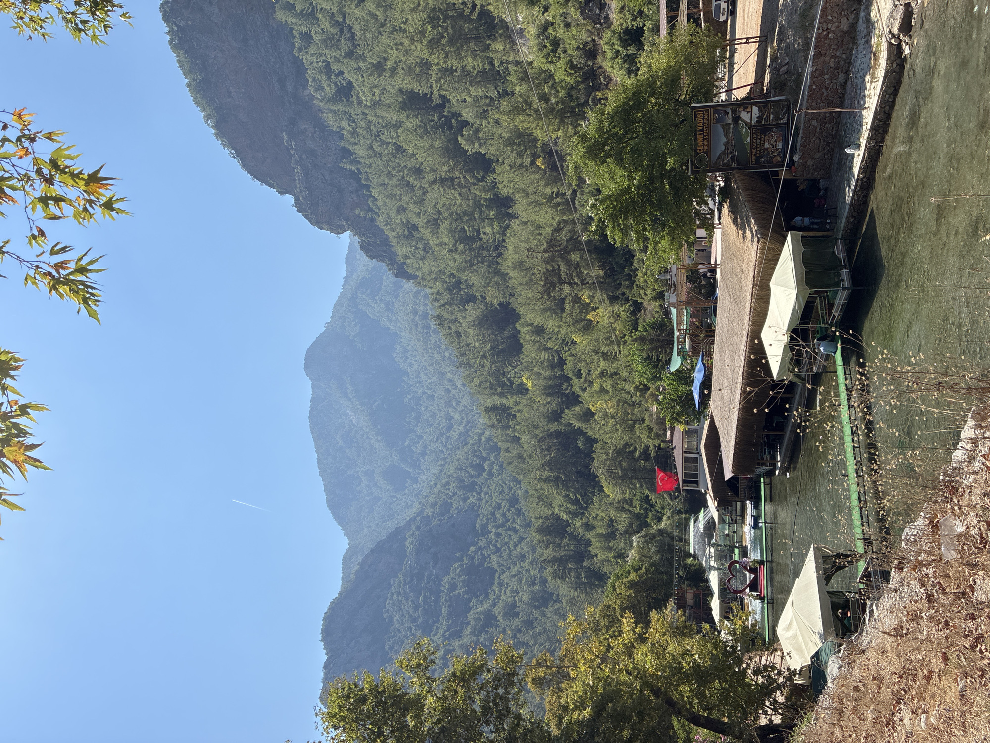 In Alanya, Turkey, a serene river scene unfolds amidst a lush, mountainous backdrop. The clear, shallow water is traversed by elevated green walkways, leading to numerous thatch-roofed dining platforms built directly over the river. These platforms are furnished with tables and chairs, some shaded by white canopies, and appear to be set up for outdoor dining. A prominent red Turkish flag flies from one of the buildings on the shore. In the distance, the mountains are densely covered in green trees, suggesting a natural, possibly forested area. The sky is a bright, clear blue, with a faint contrail visible. The foreground features dry, golden grasses, indicating a warm, sunny day.
