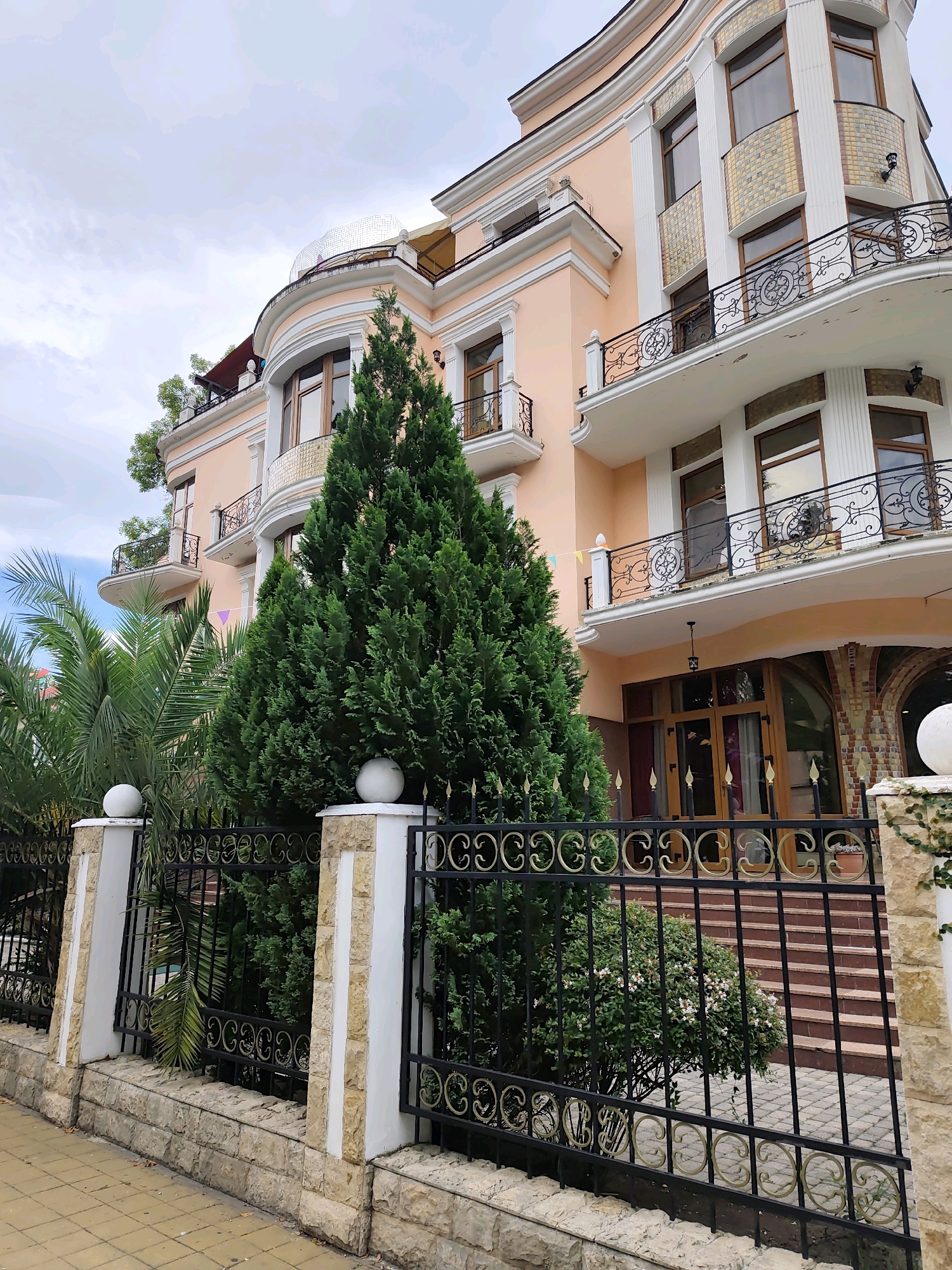 The image shows a peach-colored, multi-story building with an ornate balcony and numerous windows, set behind a black wrought-iron fence. The building has a curved facade, with some sections featuring decorative tilework around the windows and balconies. Two tall, dark green evergreen trees stand in front of the building, partially obscuring it. A palm tree is visible to the left of the evergreen trees. The fence is supported by stone pillars, some topped with white spheres. The ground in front of the fence is paved with yellow square tiles. The sky is overcast with light clouds. This scene is located on a street in Sochi, Russia. There are no people or animals visible in the image.