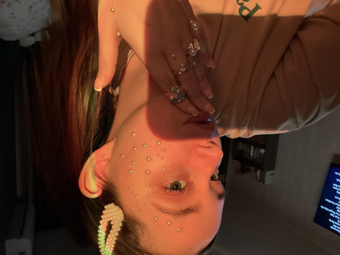 A fair-skinned person poses with their head tilted. They are adorned with small, pearl-like embellishments on their face. Their fingers, laden with rings, are positioned near their mouth. A pearl-decorated hair clip adorns their hair near their ear. The person wears a light-colored sweatshirt. A television displaying text is visible in the background. The scene is lit with an orange hue, possibly from a light source.