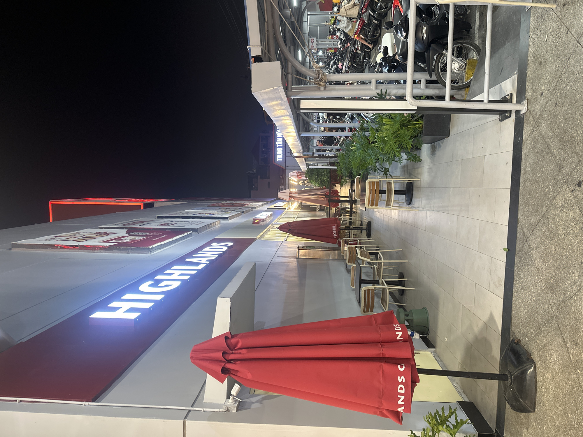 The image captures a nighttime scene outside a "Highlands" coffee shop in Nha Trang, Vietnam. The shop's illuminated sign is prominently displayed. In the foreground, a large, closed red umbrella stands on the tiled walkway. Several tables and chairs are set up outside the shop, some with umbrellas. Beyond the seating area, a parking lot is visible, filled with numerous motorcycles. A few people, some wearing face masks and helmets, are seen near the motorcycles. The environment appears to be an urban commercial area with covered walkways and bright lighting.