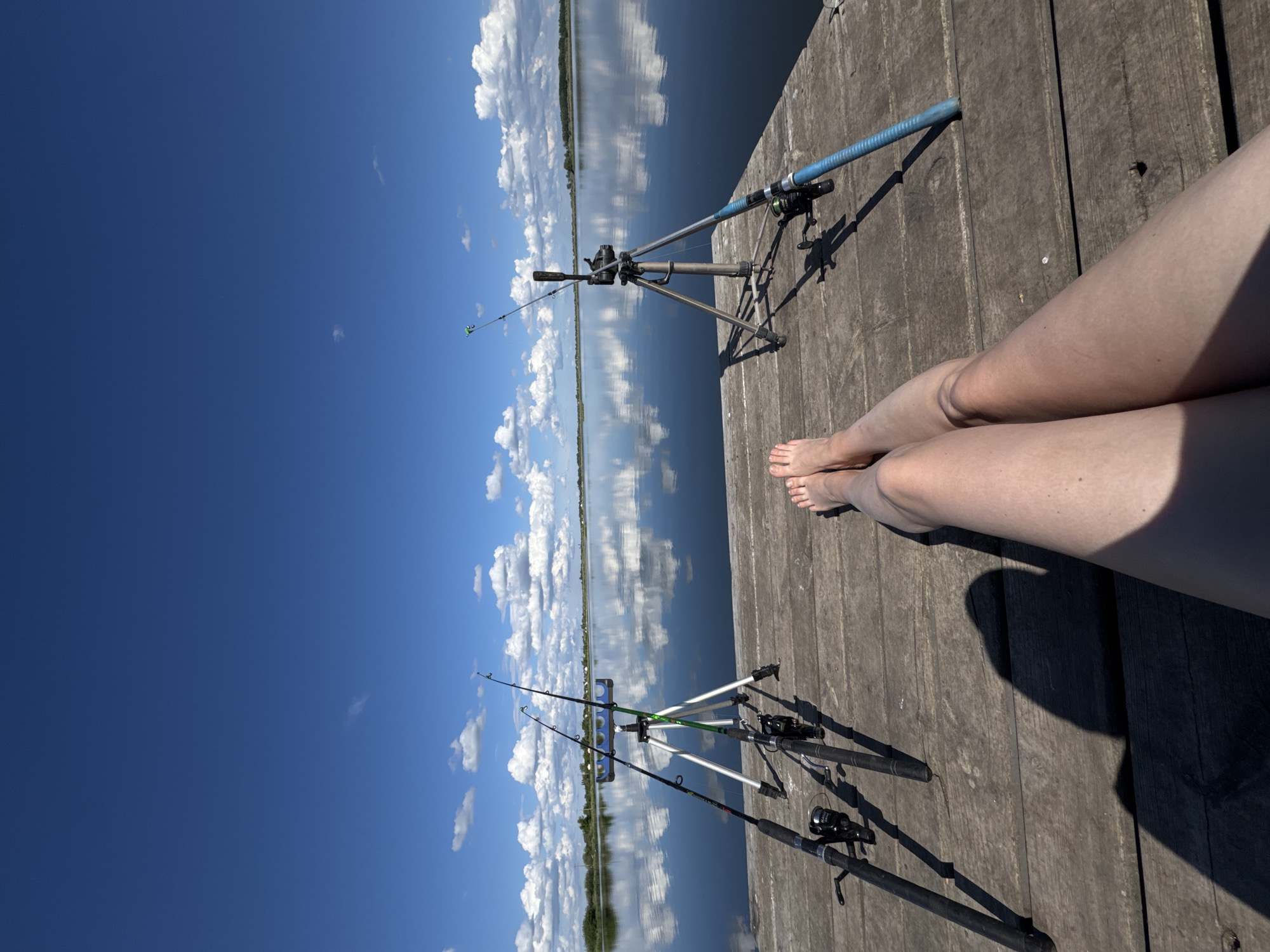 A low-angle, outdoor shot depicts a wooden dock, possibly at an Unknown, Unknown location. On the dock, there are two fishing rod holders with fishing rods inserted. A pair of bare legs extends into the frame from the upper right corner. The background features a body of water reflecting a blue sky with white clouds.