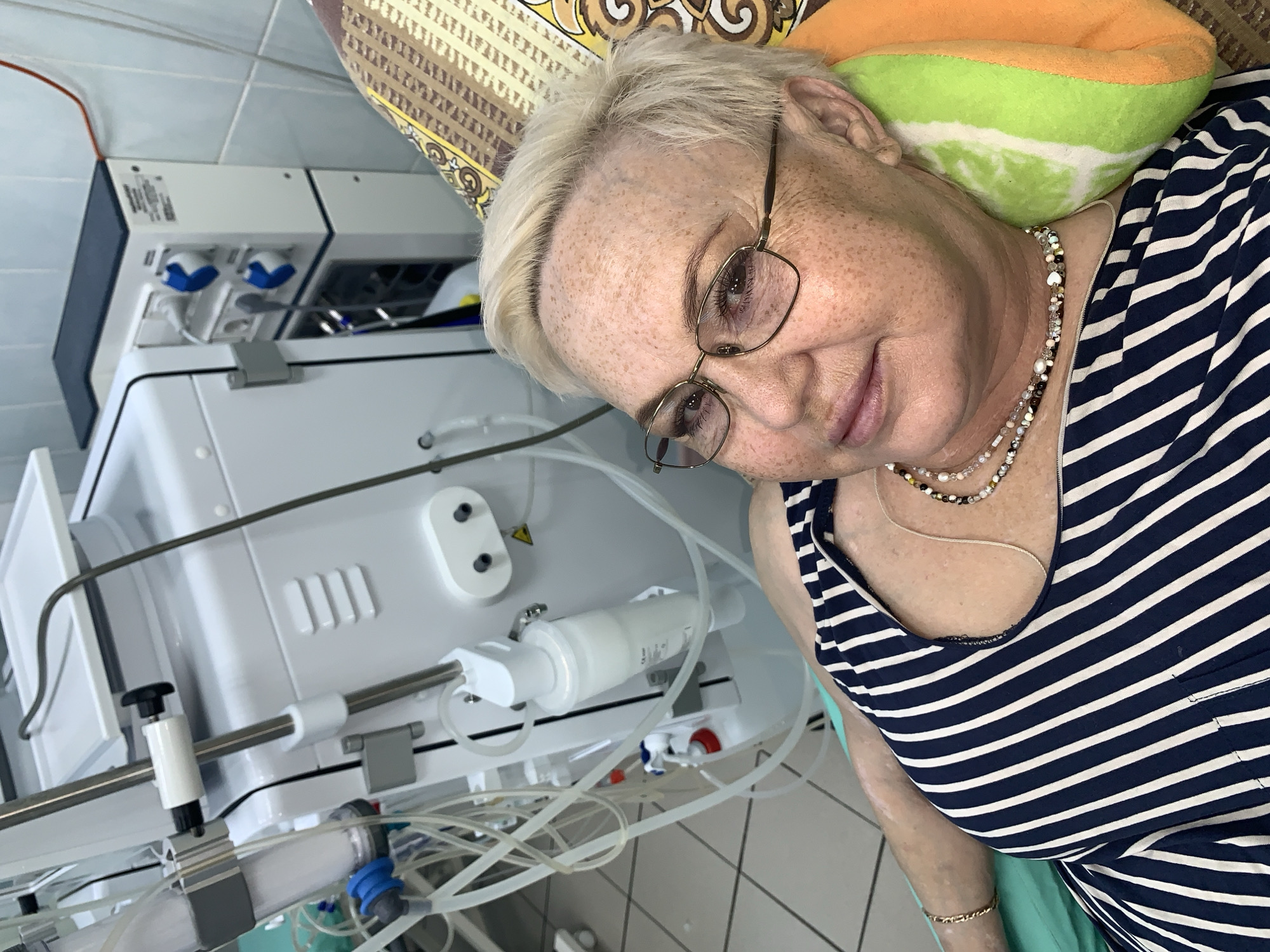 A middle-aged woman with short blonde hair and eyeglasses, adorned with a layered necklace, lies on a pillow while wearing a navy and white striped top. Beside her is a white medical machine with tubes and attachments. The scene is in the city of Chaikovsky, Russia.