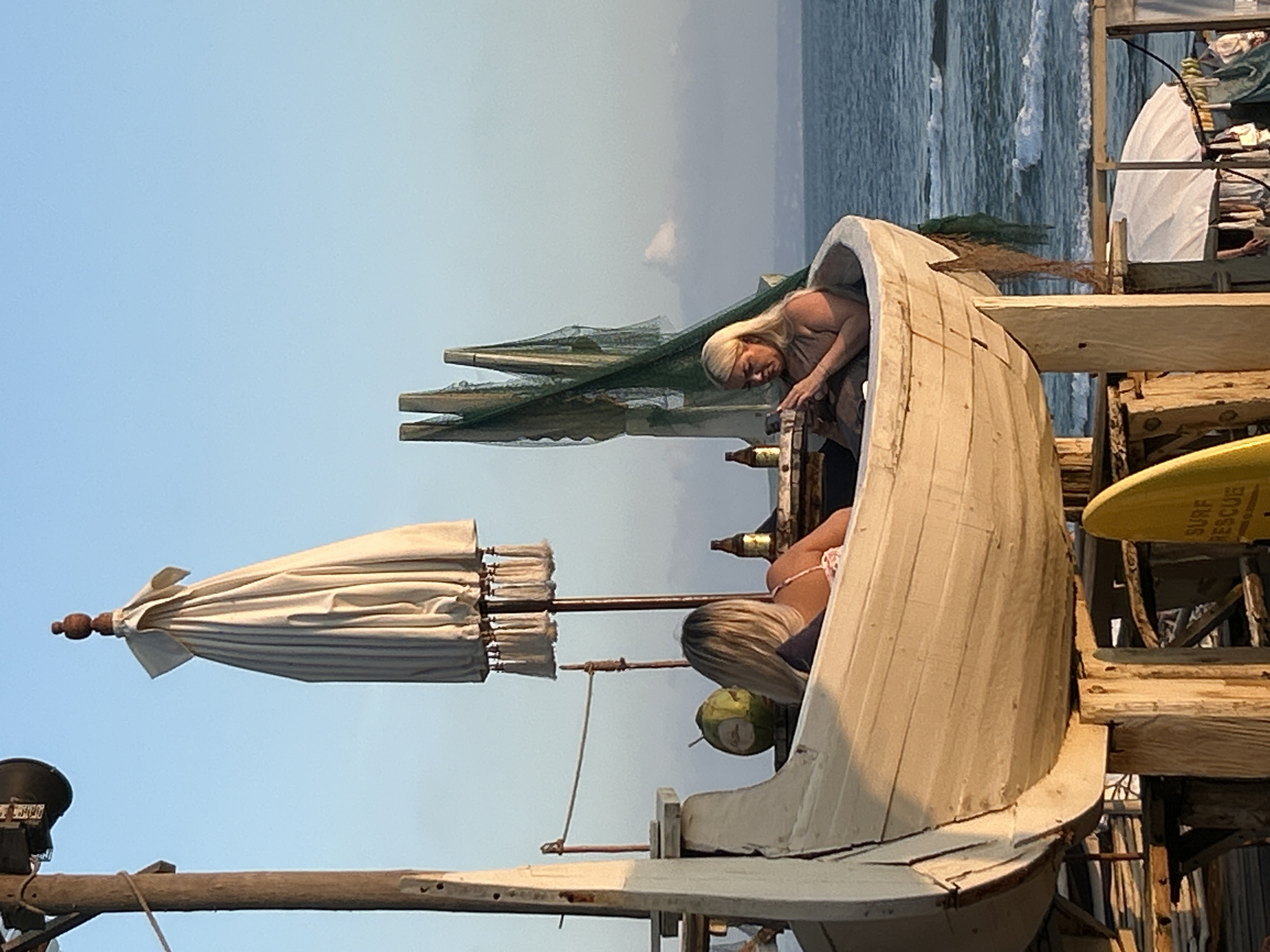 The image depicts a seaside scene at sunset in North Kuta, Indonesia. Two women are seated inside a white, boat-shaped structure supported by wooden posts. The woman on the left, with blonde hair, faces away from the camera, holding a coconut. The woman on the right, also blonde, faces left, looking down at her phone. Both women are wearing casual clothing. Behind them, fishing nets hang, and a closed white umbrella is visible. Bottles of beer and a coconut are on a surface within the boat. The ocean stretches out in the background, with gentle waves and a hazy horizon. A yellow surfboard with "SURF RESCUE" written on it is leaning against the structure in the foreground. The overall ambiance suggests a relaxed, tropical setting.