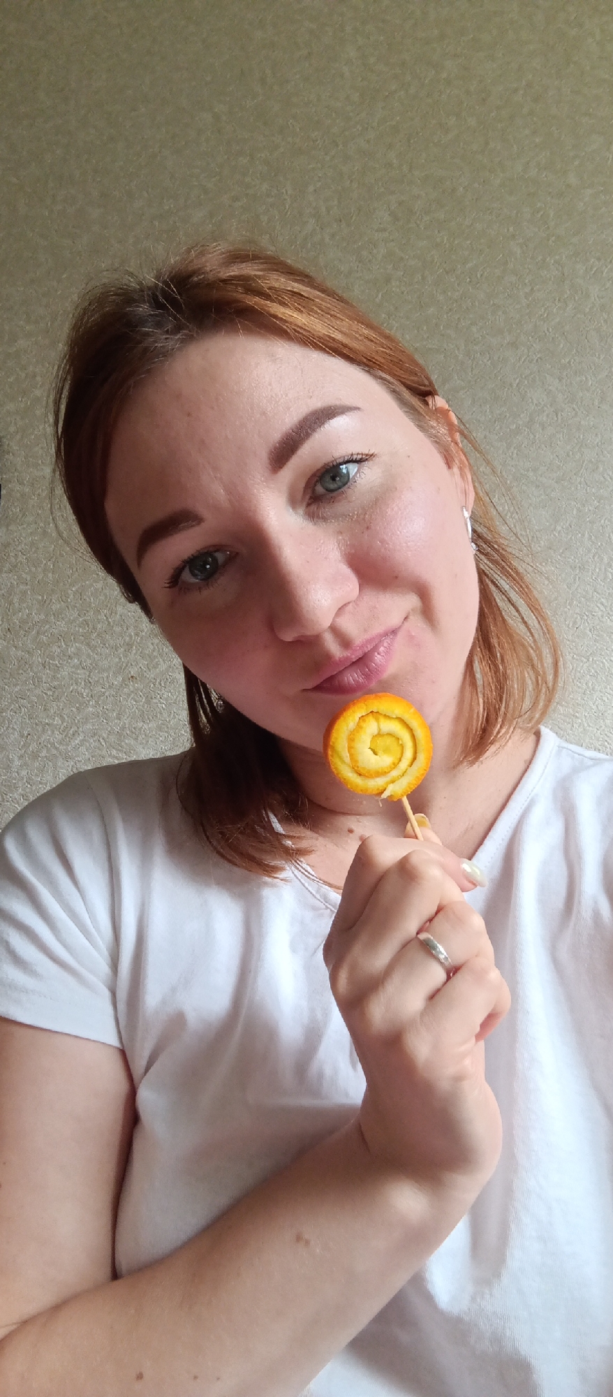 A woman with auburn hair and blue eyes is holding an orange peel lollipop in her right hand. She is wearing a plain white t-shirt and a silver ring on her ring finger. The background is a light beige wall. The location is indoors.