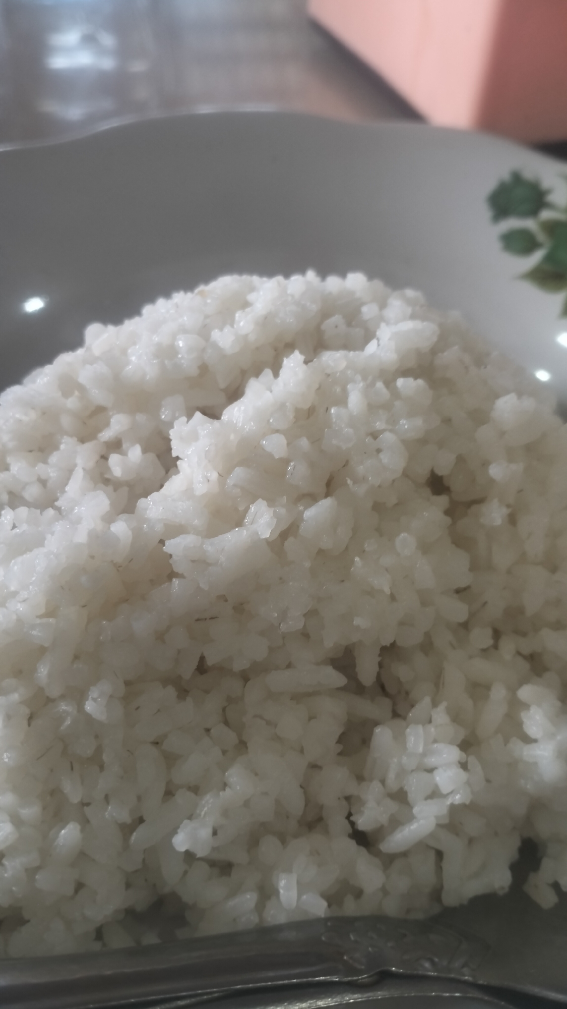 A close-up shot captures a generous portion of cooked white rice served in a white, shallow bowl or plate. The rice fills the majority of the frame, showcasing its individual, slightly translucent grains, suggesting it is freshly prepared and moist.
The plate features a subtle green floral or leafy pattern on its rim, partially visible on the right side of the image. At the bottom of the frame, a metallic eating utensil, likely a spoon or fork, is partially visible, resting against the plate. The handle of the utensil appears to have some embossed decorative details.
The background is softly blurred, indicating a shallow depth of field, and shows indistinct shapes and light colors, possibly suggesting an indoor setting like a dining table. There are no people visible in the frame, and no discernible activity other than the implied act of preparing or consuming a meal.
The image portrays a simple, staple food item often consumed in Indonesia, consistent with the provided location context of Lhokseumawe. No text or specific time of day cues are visible.