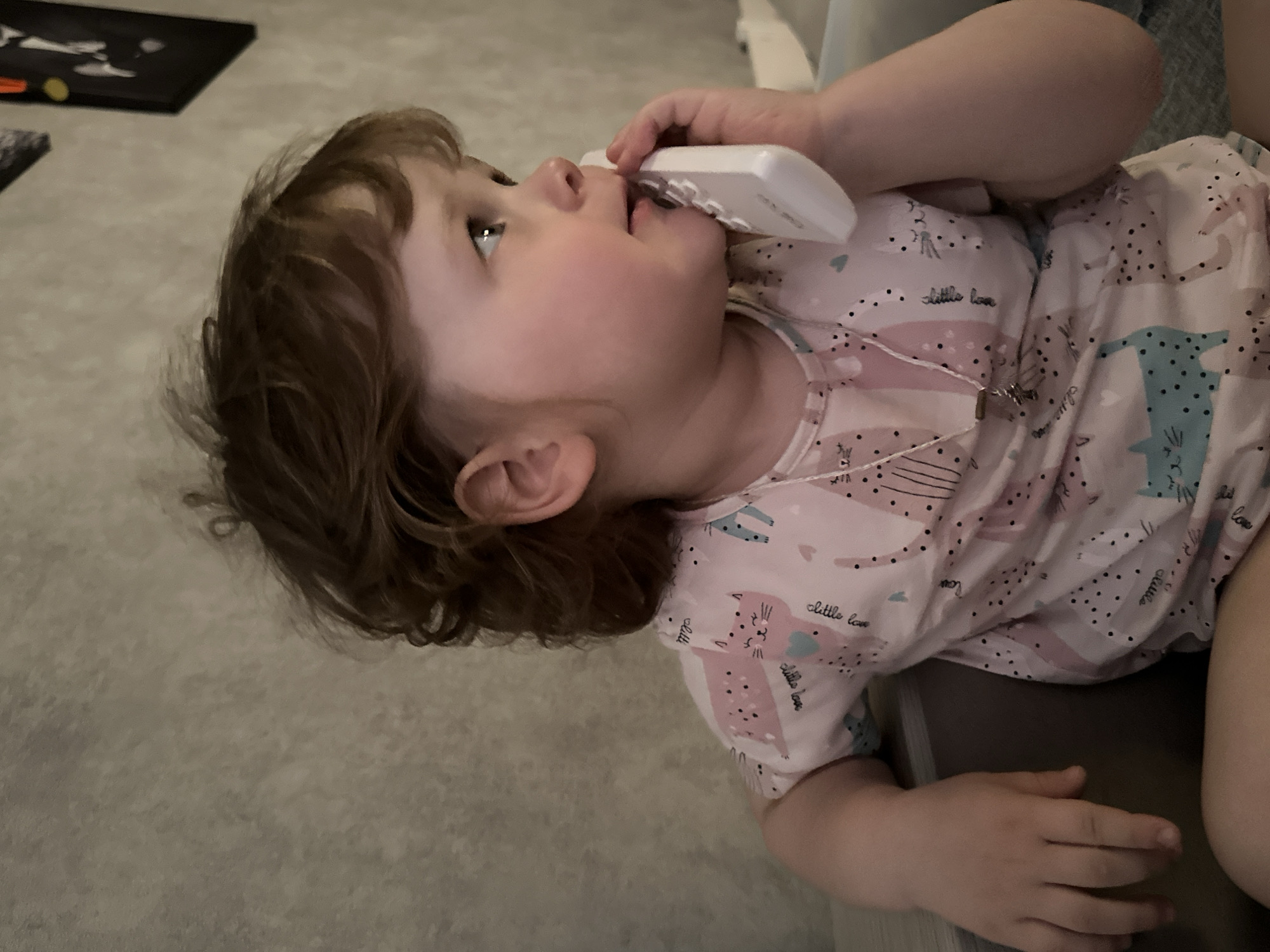 A young child with brown, curly hair and fair skin is holding a white cordless phone to her face as if talking on it. She is wearing a pink and white shirt with a cartoon cat pattern and a necklace with a pendant. The child is reclining, with her head tilted back. Her right hand rests on her chest. The background consists of a gray carpet or floor and a dark object in the upper left corner. There is also a suggestion of a bed or sofa in the upper right of the image. The location in the city of Chaikovsky, Russia is not discernible from this image.