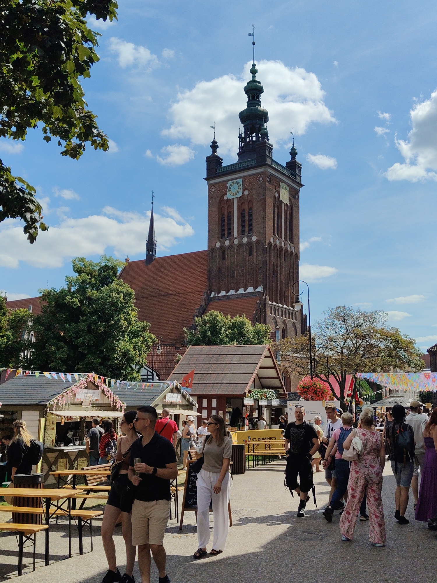 The image captures a bustling street scene with a prominent brick tower, identified as part of St. Mary's Church in Gdańsk, Poland, dominating the background. The tower features a dark green spire, a clock face, and intricate architectural details. In the foreground, a street market is in full swing, with various stalls adorned with colorful flags and decorations. Numerous people are browsing the stalls, walking along the cobblestone street, and socializing. Some individuals are seen with drinks in hand. The atmosphere appears lively and festive, with a bright, sunny day contributing to the pleasant ambiance. Trees and foliage are visible, adding to the scenic environment of the city.