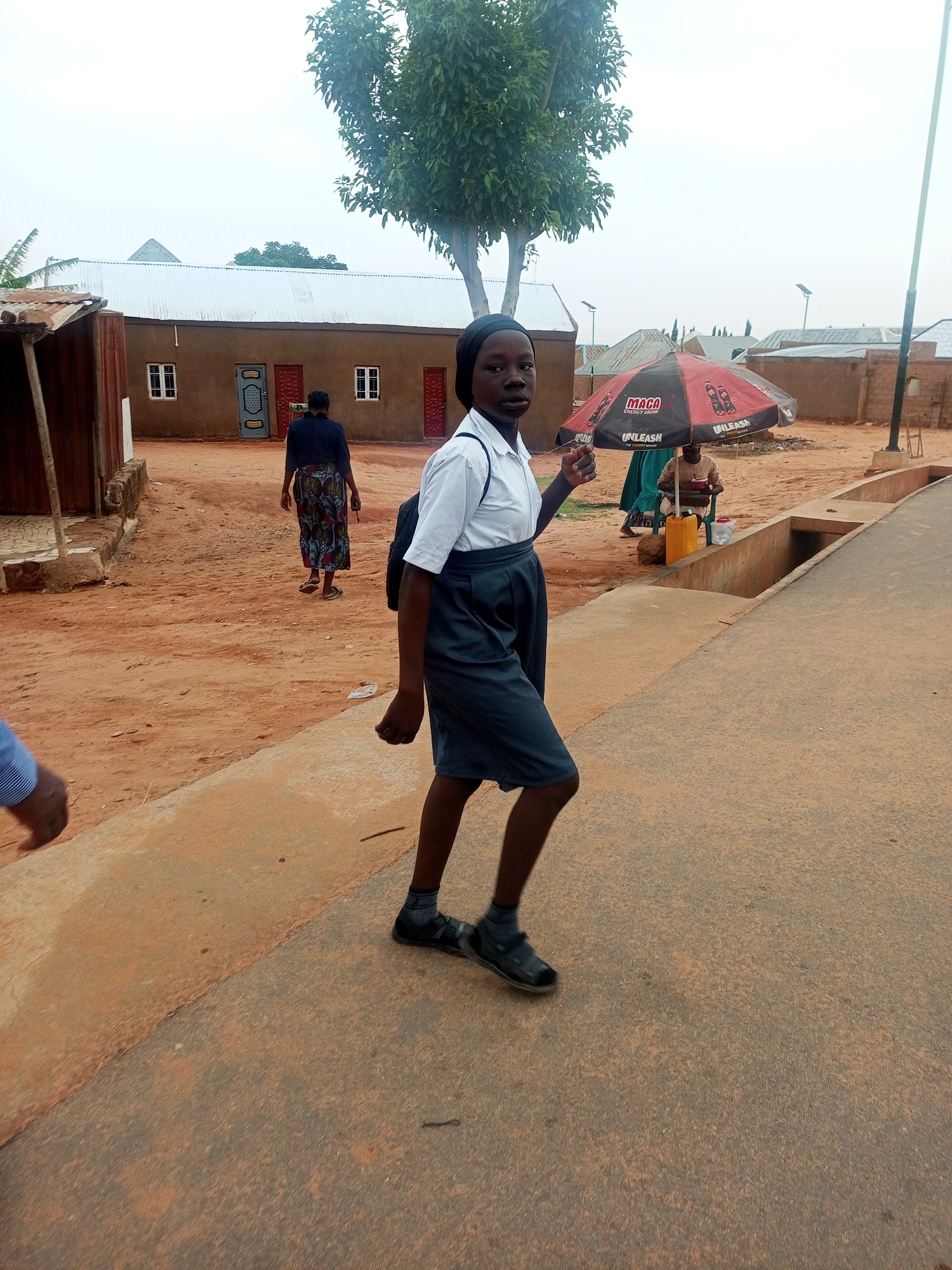 The image depicts a street scene in Tempure, Nigeria. A young woman in a school uniform walks along a concrete road. In the background, another woman walks away from the camera towards a mud-brick building with a corrugated metal roof. Under a red and black umbrella, a person sits on a stool by a yellow container. The sky is overcast, and the surrounding environment appears to be a residential area with dirt roads and simple structures. A tree is visible in the middle ground.