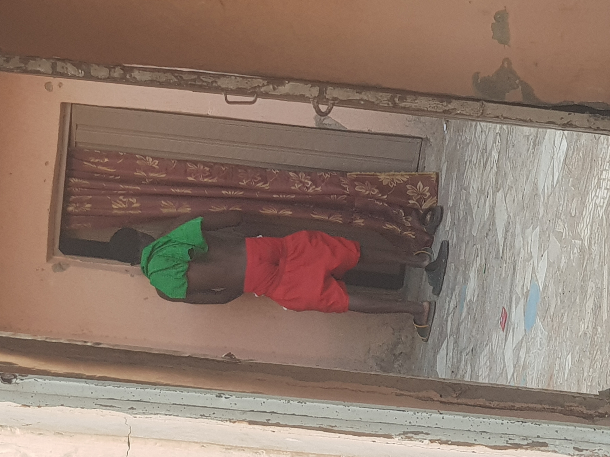 A person wearing red shorts and a green head covering is inside a doorway of a building with light peach walls. The doorway has a metal roll-up door partially lowered, behind which a patterned curtain is visible. The person is wearing sandals. The floor is composed of patterned tiles. The location is in a city, potentially Dutsinma A, Nigeria.