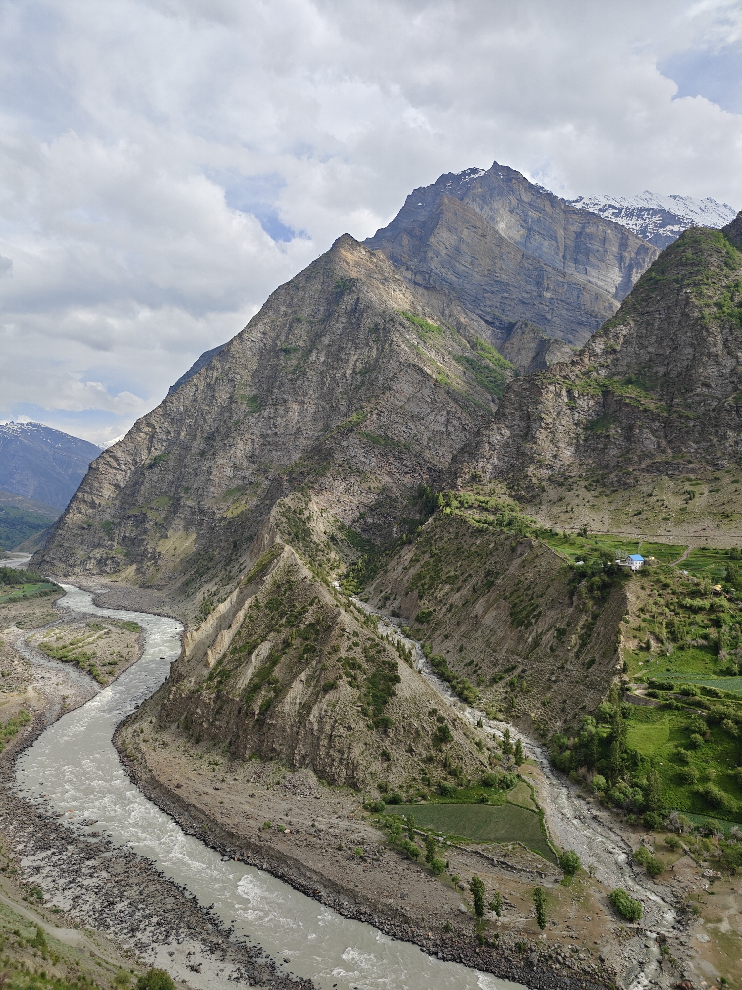 This aerial, high-angle, medium shot captures a majestic mountain landscape in the region of Kirting, India. The scene is dominated by rugged, steep mountainsides with varying textures of rock and sparse vegetation. A wide, turbulent river flows through the valley, its milky, silt-laden waters carving a winding path through the rocky terrain. The riverbanks are a mix of stone, gravel, and patches of green vegetation, indicating an arid to semi-arid climate.
In the middle ground, a solitary house with a bright blue roof stands on a terraced hillside, surrounded by small, neatly cultivated green fields. The architecture suggests a rural dwelling adapted to the mountainous environment. The surrounding slopes are dotted with scattered trees and shrubs, with patches of green fields visible on the higher terraces, hinting at human cultivation in this remote area.
The background features progressively larger and more imposing mountains, with the peaks of some of the highest mountains dusted with snow, suggesting significant altitude. The sky is filled with a dramatic mix of clouds, ranging from light wisps to dense, grey formations, indicating dynamic weather conditions. The overall impression is one of a vast, untamed natural environment interspersed with signs of human habitation and resilience. There are no people or animals visible in the frame.