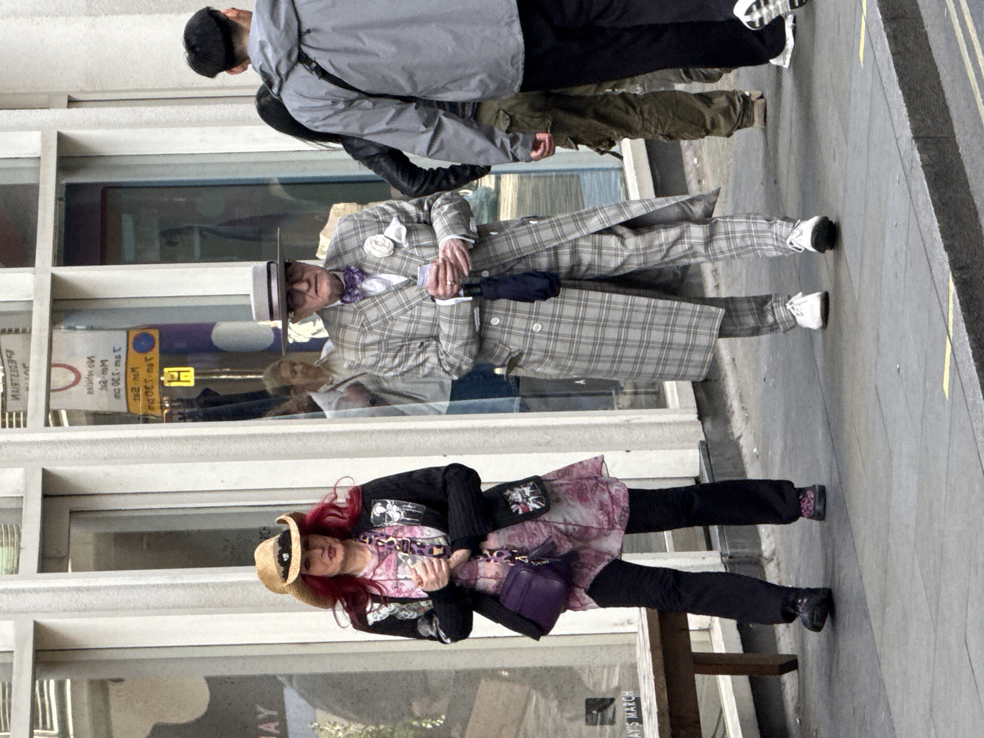 The photograph shows a street scene in what appears to be London, United Kingdom. Three pedestrians are walking on the sidewalk.
In the foreground, a woman with red hair wearing a straw hat, a patterned pink dress, a black jacket, and black pants walks facing forward with a purple handbag. Behind her stands a man wearing a plaid suit, a matching hat, and white sneakers. He is holding a black umbrella. A gray jacketed person with dark pants can be seen walking behind him.
The pedestrians are walking in front of a building with large windows. Reflections are visible in the glass.
Based on the visible signage “Sloane Street” in the reflection and “AY'S MARCH,” the location may be near Sloane Street.