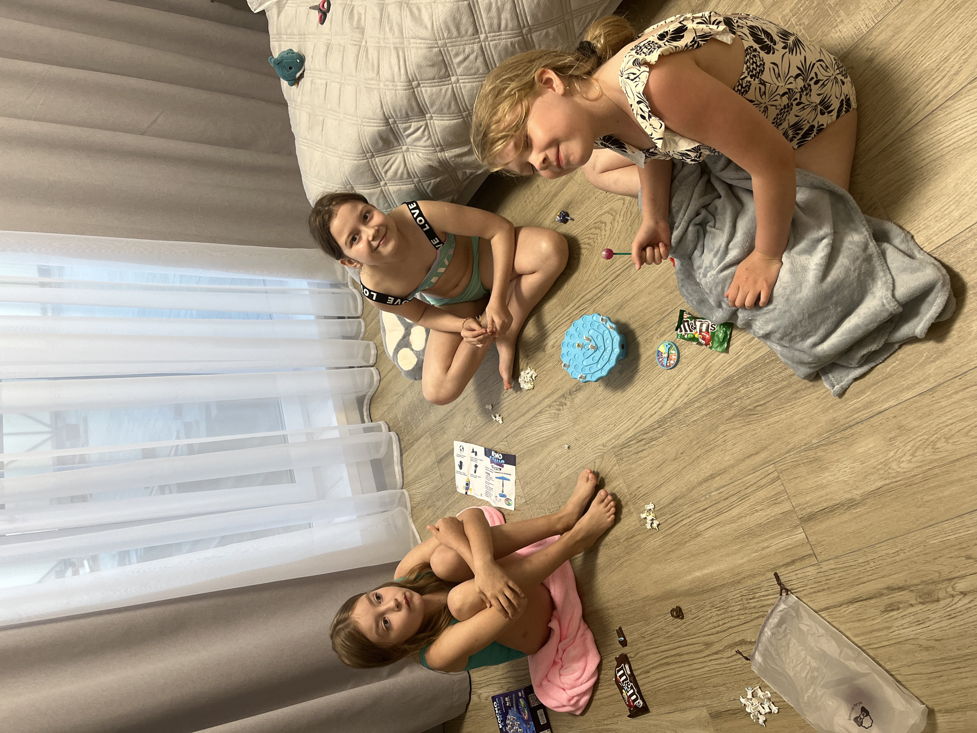 Three young girls are on the floor of a room in Новая Чернушка, Russia. The room contains a bed with a grey patterned comforter, curtains, and a wood-look floor. The girl closest to the camera is seated on a pink towel, hugging her knees. To her right is a box, candy, and a transparent bag of white toy pieces. Above her and to the right is another girl seated on a spotted pillow, wearing a two-piece swimsuit with black lettering. Next to her is a blue toy. The third girl is wearing a printed one-piece swimsuit and is partially covered with a grey blanket. Candy wrappers and small white toy pieces are scattered around the floor. The girls appear to be playing.