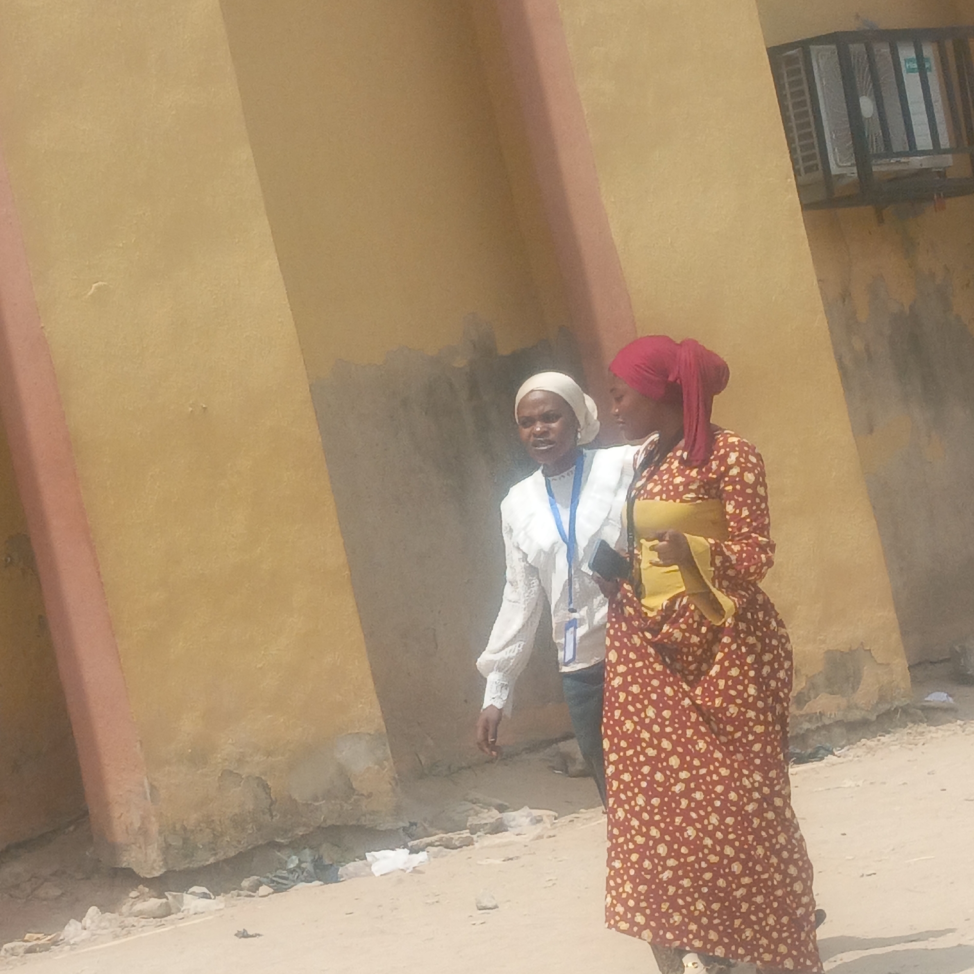 This daytime image, set in Keffi, Nigeria, features two women standing on a sandy or dirt path next to a building with a weathered, yellowish-orange wall. The bright sunlight suggests a clear day.
The woman on the left wears a white head covering and a white long-sleeved blouse with ruffled details, paired with dark blue pants. A blue lanyard with an ID card, featuring a blue frame, hangs around her neck. She appears to be speaking or engaged in conversation, with her mouth slightly open.
The woman on the right wears a red head covering and a long, flowing dress or skirt and top ensemble. The garment is red with a scattered pattern of small, light-colored (possibly white or light yellow) floral or polka dot designs. A plain yellow top is visible underneath her outer layer. She holds a dark mobile phone in her left hand and is looking downwards.
The building wall behind them shows considerable wear, with peeling paint, discoloration, and dark stains, particularly near the base. Prominent reddish-brown vertical architectural elements or pillars are visible on the left side and partially behind the women. In the upper right corner, an air conditioner unit, possibly white and green, is mounted in a window and protected by a dark metal grille or cage. Faintly visible text on the air conditioner unit includes characters that appear to be "HE" or "HL". The ground is dry, light-colored soil or sand, with scattered debris and small pieces of litter.