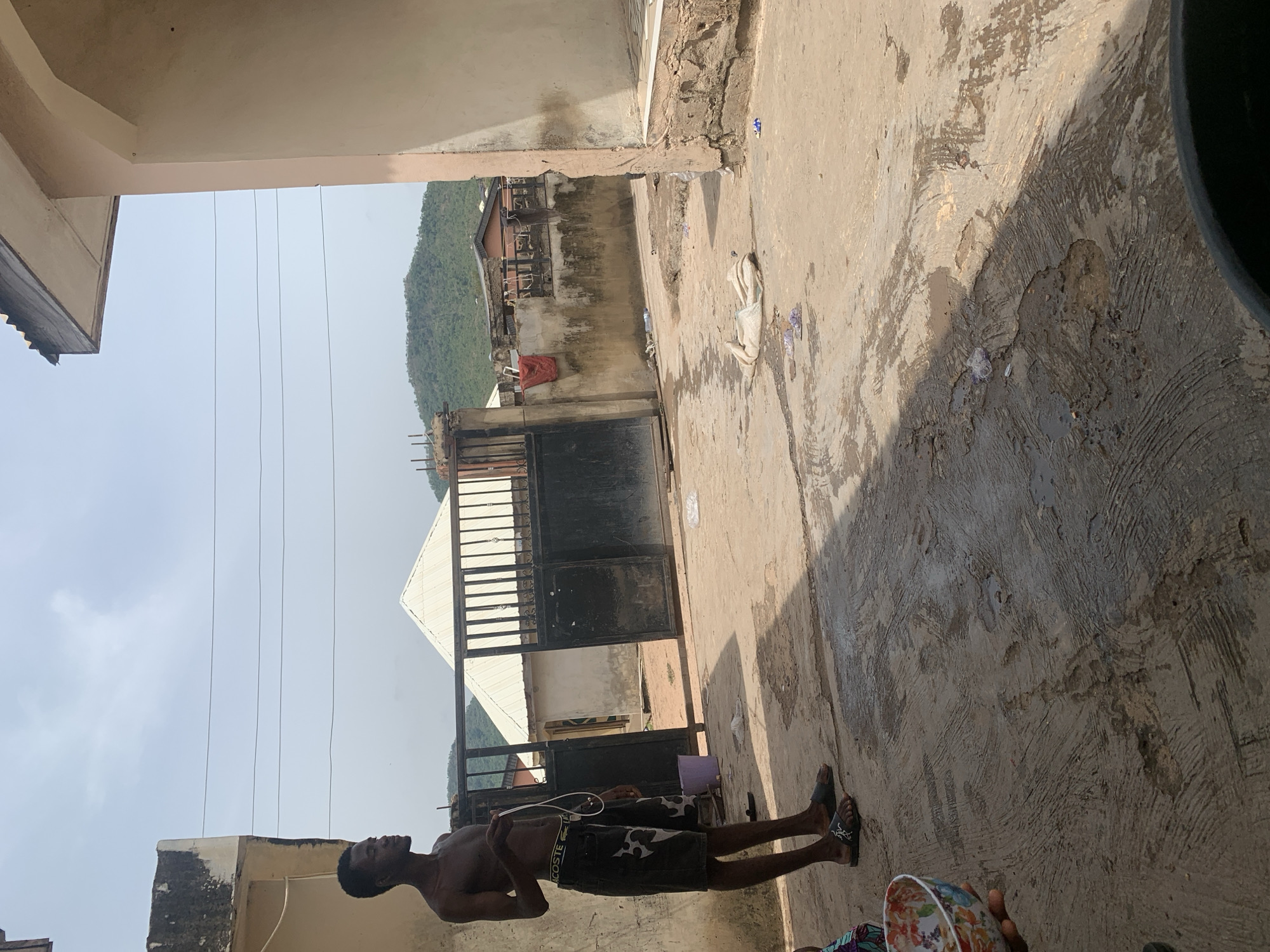 The image shows a view from a concrete-surfaced courtyard or alleyway, potentially in Lokoja, Nigeria. A shirtless man wearing dark shorts stands on the left side of the frame, holding what appears to be a white cord or strap. He faces the open area of the courtyard.
In the background, there is a gated entrance or fence, beyond which a white-roofed structure is visible. Behind the structure, there is a distant view of a hill covered in green vegetation.
The surrounding buildings are constructed of concrete and appear to be in a state of disrepair. There are electrical wires visible overhead. The ground is uneven, with visible signs of wear and discoloration. In the bottom right, there is part of a bowl.