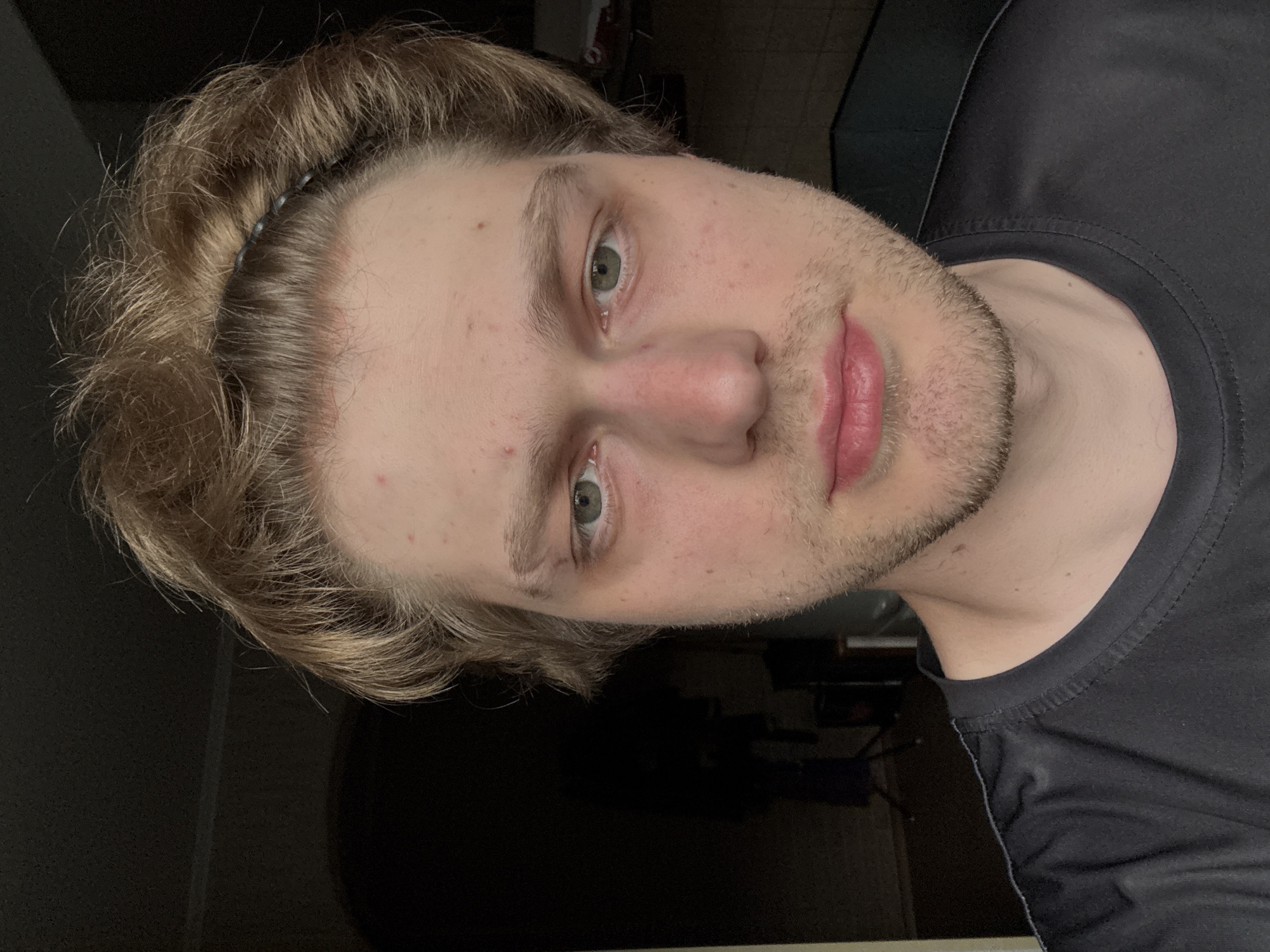The image is a close-up, vertically oriented selfie of a young man in Khimki, Russia. He has light brown hair, styled with a dark headband, and fair skin with some minor blemishes. His eyes are green, and he has light stubble on his face. He is wearing a black t-shirt. The background is dark and indistinct, suggesting an indoor setting, possibly at night or in a dimly lit room. There is no discernible weather or time of day information, other than the implication of it being indoors. The man's expression is neutral. There is no visible text in the image.