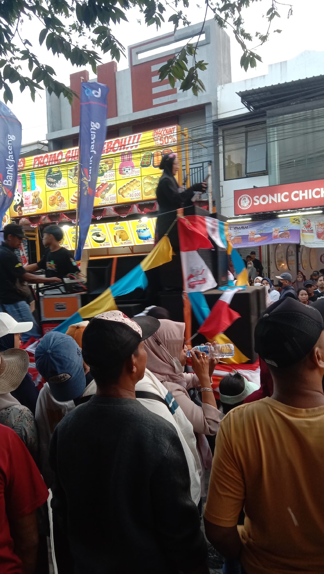This image captures a street scene in Jatisawit, Indonesia, likely during a promotional event or festival. In the foreground, the backs of several people are visible, dressed in casual attire. One person in the center wears a dark grey sweatshirt, while another to the right is in a yellow t-shirt. In the middle ground, a group of people are gathered around a stage or booth, where a person in dark clothing appears to be speaking or performing, holding a microphone. Colorful flags and banners are displayed, some featuring text that is partially obscured but appears to include "Bank Jateng" and promotional offers like "PROMO SPESIAL" and "DISCOUNT UP TO 50%". Several food items are advertised on a display board with prices indicated in Rupiah, such as "Rp 45.000" and "Rp 25.000". The background shows buildings with a mix of modern and traditional architecture, with some overhead wires suggesting an urban environment. The overall atmosphere suggests a lively community gathering.