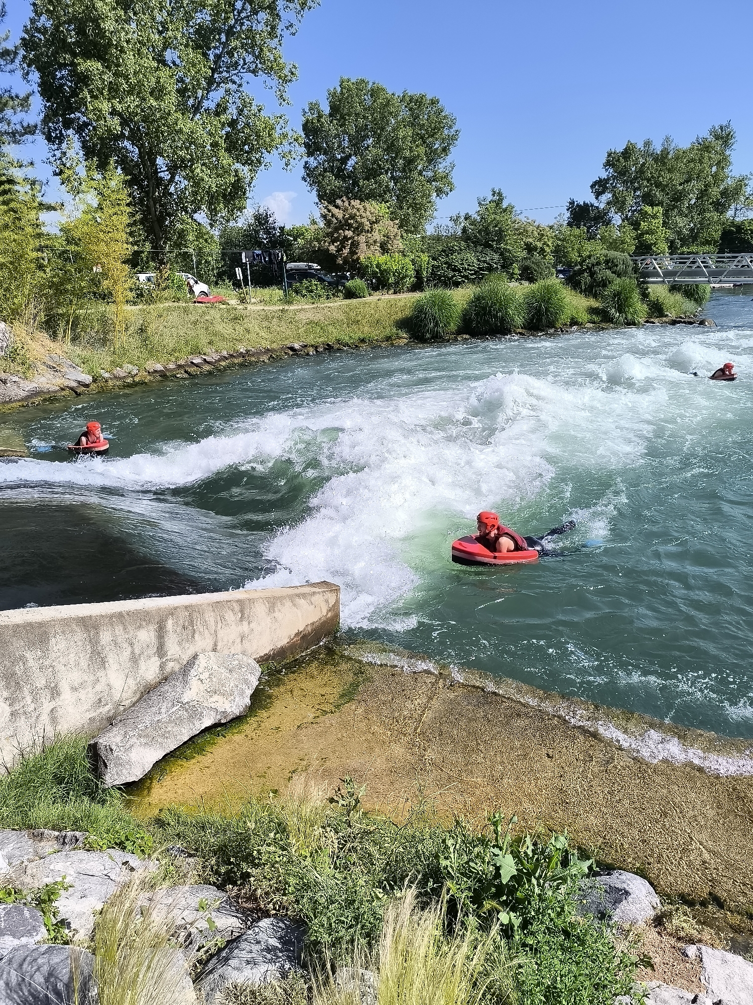 The image shows three people bodyboarding on the artificial whitewater course at Saint-Pierre-de-Bœuf, France. The location is identifiable by the prominent concrete structures and landscaping along the artificial course and the bridge in the background.
In the foreground, a person wearing a red helmet, a black vest, and fins is riding a red bodyboard in turbulent water near a concrete structure. Another person is further up the channel, while a third is visible in the distance. The water is a light green-blue color, with areas of white from the rapids.
The shoreline is lined with green vegetation, trees, and grass. In the background, there are more trees, a glimpse of a building, and a metal bridge spanning the water. The sky is clear and blue.
