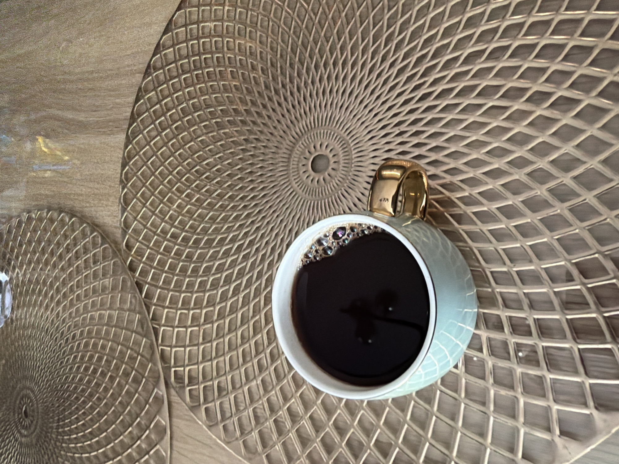 An overhead shot captures a light blue ceramic coffee mug with a golden handle, filled with dark coffee, resting on a golden, diamond-patterned placemat. To the left of the mug, another identical placemat is partially visible. The scene is set on a wooden table. No people or specific locations in Rafailovići, Montenegro, are depicted.