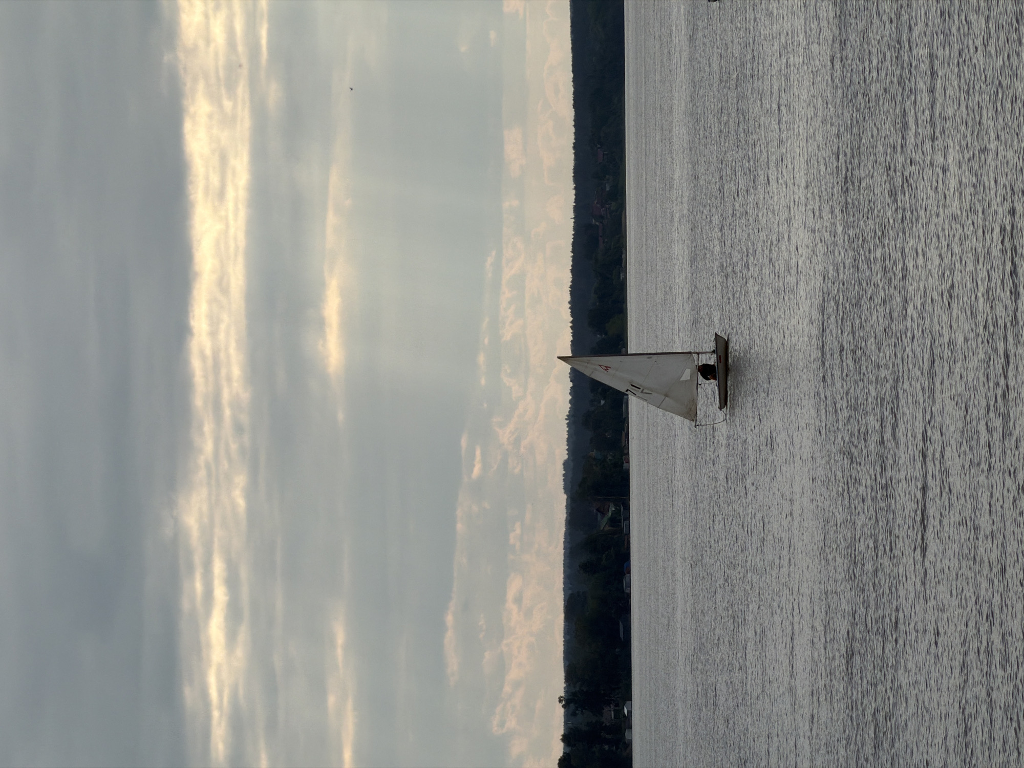A sailboat with a single occupant is sailing on a body of water. The sailboat has a white sail with red markings. The occupant appears to be sitting in the boat. The water is calm with small ripples. In the background, there is a tree-lined shore with some buildings visible amongst the trees. The sky is cloudy with shafts of sunlight breaking through. The image was taken from a distance, suggesting an elevated viewpoint or a zoomed-out perspective. The overall scene depicts a peaceful day on the water. The specific location in Votkinsk, Russia is not determinable from the image alone.