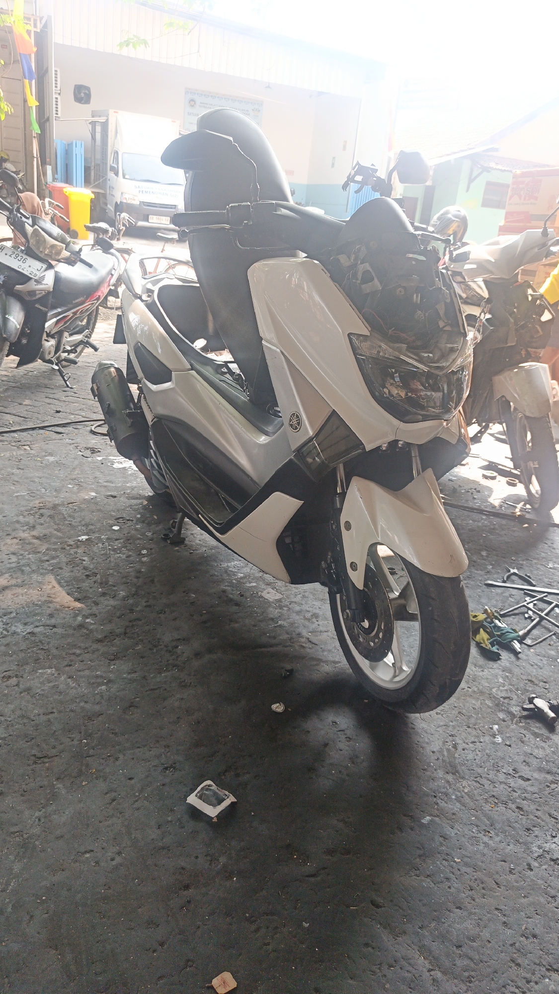The image depicts an outdoor scene in what appears to be a workshop or service area in Mlati Kidul, Indonesia. The foreground is dominated by a white scooter, likely a Yamaha NMAX, parked on a concrete floor. The scooter is the primary subject, with its front wheel and part of its body visible. Other motorcycles are also present in the background, suggesting a commercial establishment related to vehicles. A white van is visible in the background, along with some signage that might indicate the name of the business. The environment is somewhat cluttered, with tools and various items scattered on the ground, indicative of an active workspace. The lighting suggests it is daytime, with bright sunlight casting shadows and creating a glare in some areas of the image. There are no people clearly visible in the frame.