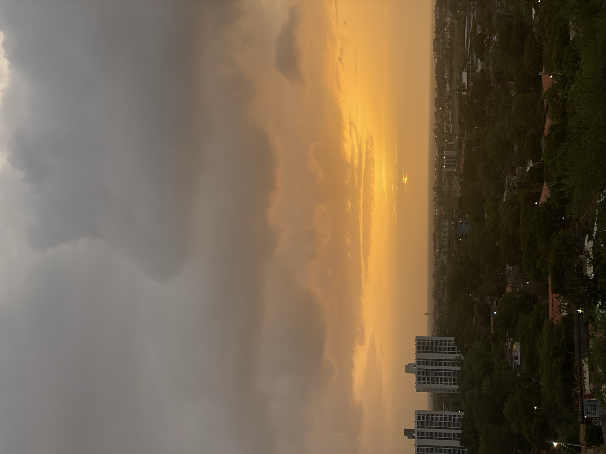 This is an aerial, horizontal shot capturing the coastal city of Juan Griego, Venezuela during a dramatic sunset. The sky is dominated by thick, dark gray clouds in the upper portion, transitioning to vibrant hues of orange and yellow near the horizon, where the sun is partially obscured. Below the sky, the cityscape is visible, featuring several tall, white apartment buildings on the left, interspersed with dense green foliage and smaller structures. The landscape extends to the distant coastline, where the sea is barely discernible against the twilight. The overall impression is one of a moody and atmospheric end to the day in a tropical urban environment.