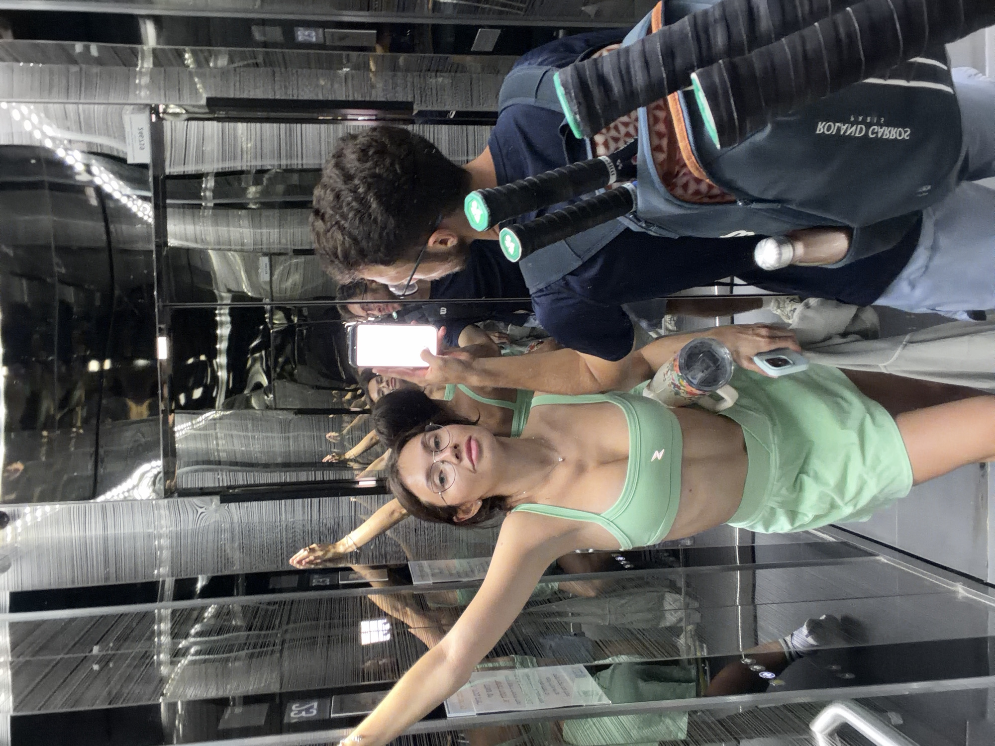 A high-angle medium shot depicts a man and a woman, their reflection visible in the mirrored elevator walls. The man, with short, dark curly hair, wears glasses and a navy blue shirt. Over his shoulder is a tennis racquet bag. He holds up a white smartphone in his right hand, as if taking a photo. The woman, with long dark hair and glasses, is wearing a light green two-piece athletic outfit. She extends her arm toward the mirrored wall, hand slightly open. The location is an elevator.