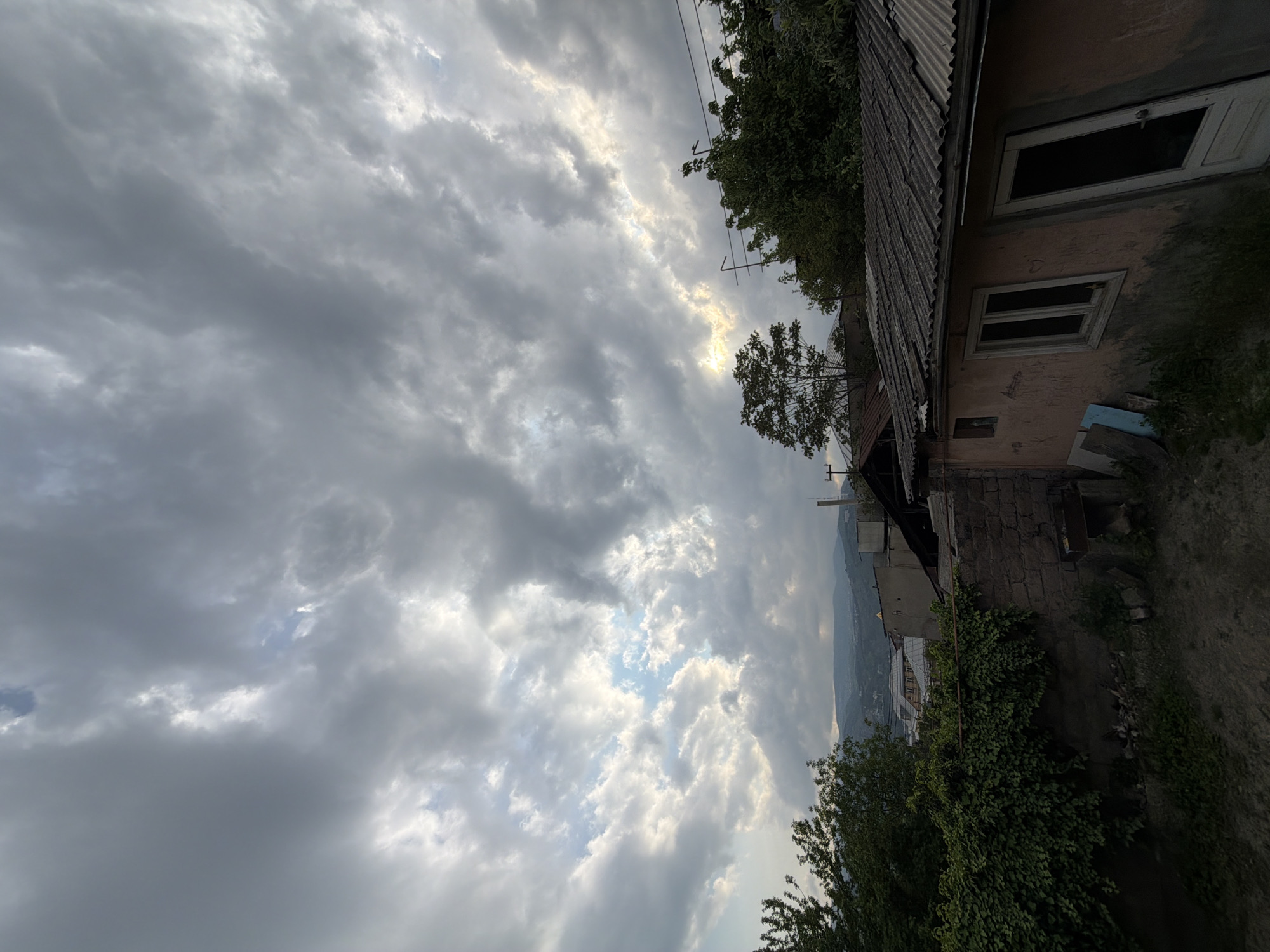 The image presents a view of a cloudy sky over what appears to be a residential area, potentially in Tbilisi, Georgia. An old building with a corrugated roof stands to the right of the frame. A window and a door can be seen. The building is adjacent to a stone wall covered in vegetation. In the background, there is a glimpse of a mountain range. Overhead electrical wires are also visible.