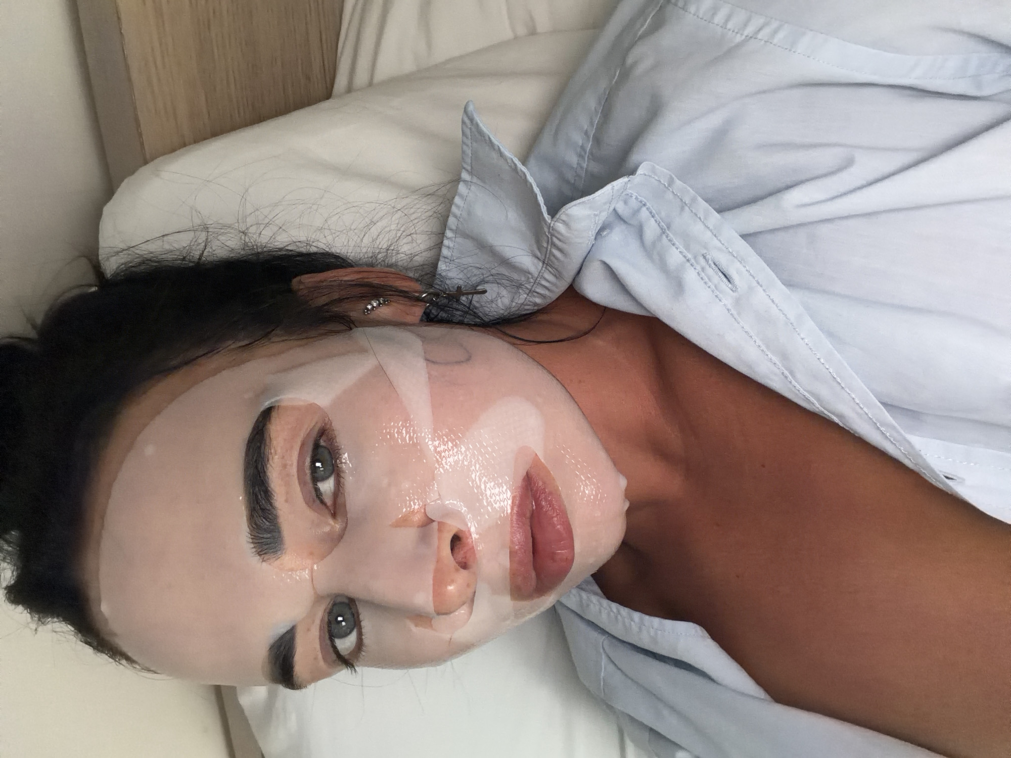 In this close-up, vertically oriented media file, a woman is lying in bed, wearing a light blue button-down shirt and a sheet mask on her face. Her eyes, with dark eyebrows and visible lashes, are open and looking towards the camera. The sheet mask covers her nose, cheeks, and chin, leaving her eyes, lips, and forehead exposed. Her lips are slightly parted. Her dark hair is visible around her head and resting on the pillow. She is wearing a small, sparkling earring on her left ear. The setting appears to be a bedroom, with a white pillow and a wooden headboard visible. The lighting suggests it could be daytime. The overall mood is relaxed, suggesting a moment of self-care or relaxation. There is no visible text in the image.