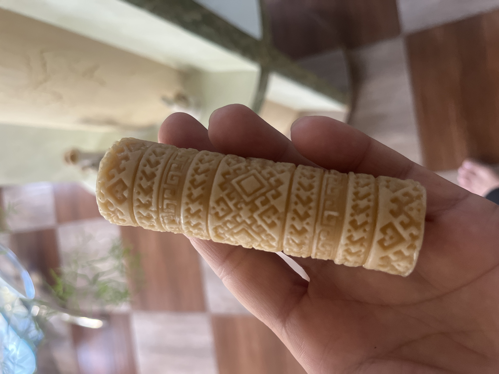A person's hand is holding a small, carved, cylindrical object with intricate geometric patterns. The object is light-colored, possibly made of bone or wood. The background is blurred but appears to be an indoor setting with a tiled floor and a light-colored structure. The image does not provide any information about the location in the city of Tarnivtsi, Ukraine.