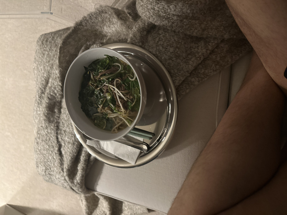 A bowl of pho sits atop a silver tray, accompanied by chopsticks and a napkin. The tray rests on a light-colored, cushioned surface, likely a sofa or ottoman. A gray knit blanket is partially visible. In the background, a portion of someone's bare legs can be seen. The scene suggests a casual meal enjoyed in a comfortable, relaxed setting.