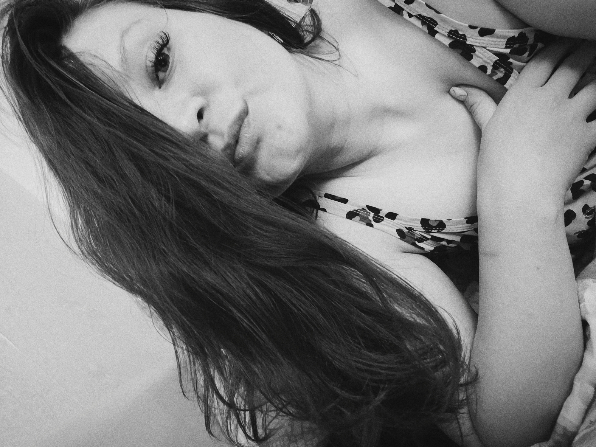 A black and white close-up shot shows a person, presumably a woman, lying down and facing the camera with a tilted head. Long, dark hair partially covers the face, particularly obscuring one eye. The visible eye has long eyelashes. The person is wearing a patterned top with a sleeveless design. The left hand is positioned near the chest area. The background is indistinct, appearing to be a flat surface, possibly a wall. No elements indicating Tobolsk, Russia are visible in the image.