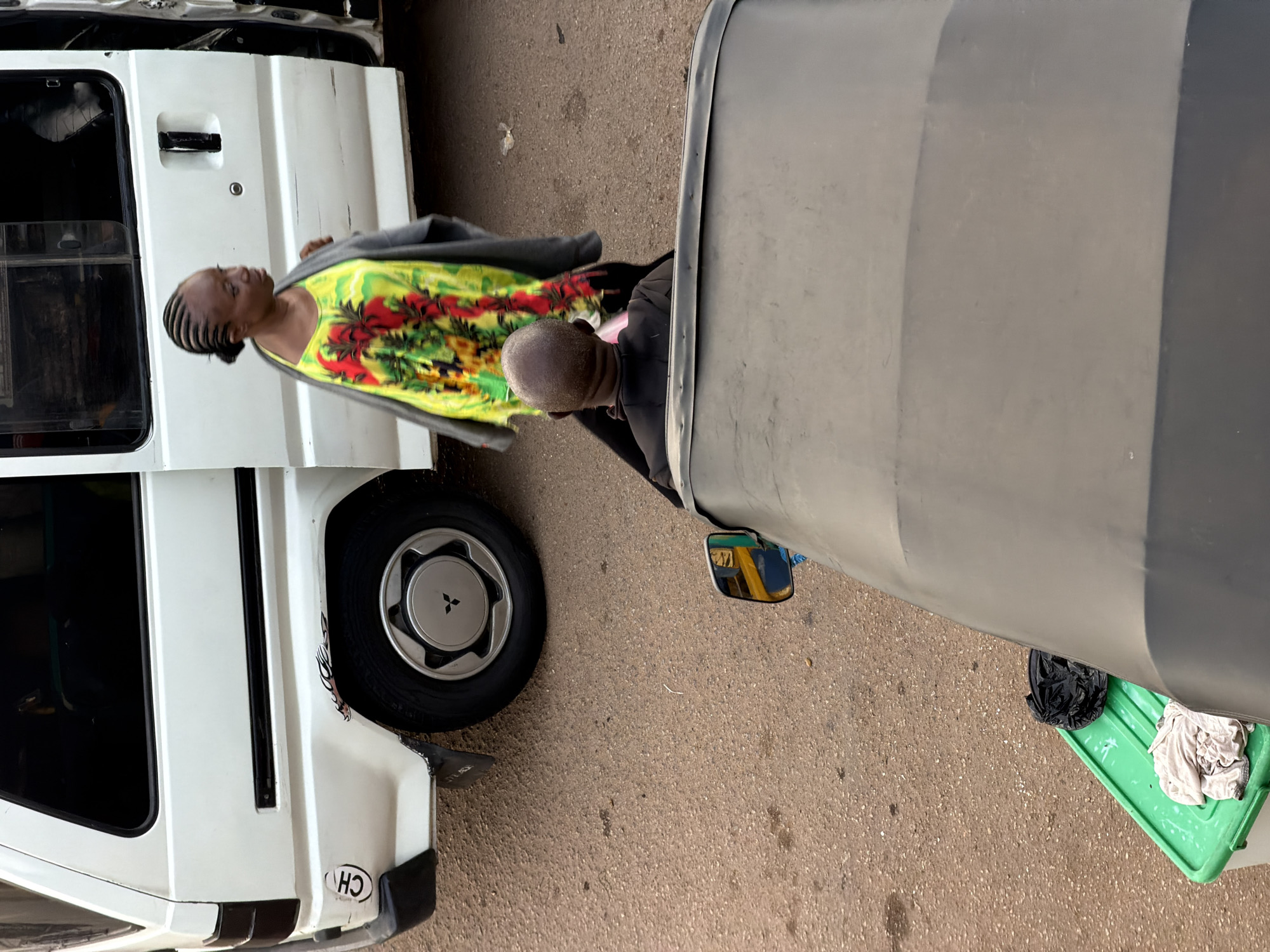 The image captures a scene on a street in Bukuru, Nigeria, with a white van and two individuals. A woman with braided hair is walking past the van, wearing a vibrant yellow, green, and red patterned dress and a grey cardigan. She is looking to her left. In the foreground, a person wearing a black shirt is seated in a dark-colored vehicle, likely an auto-rickshaw, and is facing away from the camera. Next to the van, on the ground, is a green plastic container with some light-colored fabric and a black plastic bag on top. The ground is paved with asphalt. The overall scene appears to be a street view, possibly in a market area or a busy thoroughfare.