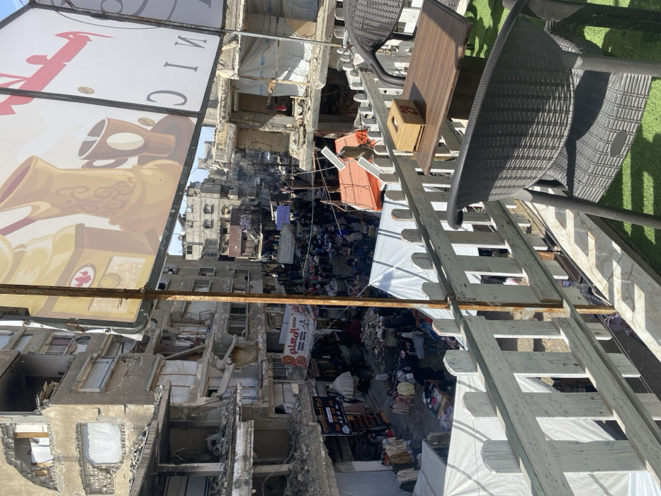 This image presents an elevated view of a bustling street market in Khan Yunis, Palestinian Territory, set against a backdrop of extensive urban destruction.
In the immediate foreground, the perspective is from a cafe or balcony featuring a white picket fence railing, green artificial turf flooring, a dark wicker-patterned chair, and a small wooden table with a wooden tissue box labeled "29." A large commercial billboard, partially visible overhead, depicts an illustrated coffee pot, a mug, and Arabic text.
The midground and background reveal a densely populated street market. Numerous individuals are visible, navigating between stalls covered by white and orange tarps, where merchandise, including clothing, is displayed on tables and racks. The street is flanked by multi-story concrete buildings, many of which exhibit severe structural damage, including collapsed sections, exposed interiors, missing walls, and visible rubble. One damaged building on the right side features a white tarp covering a section, with a partial "UNICEF" logo discernible. Further down the street, more damaged structures and a few vehicles are observed. The scene depicts active commerce occurring amidst widespread architectural devastation under daylight conditions.
