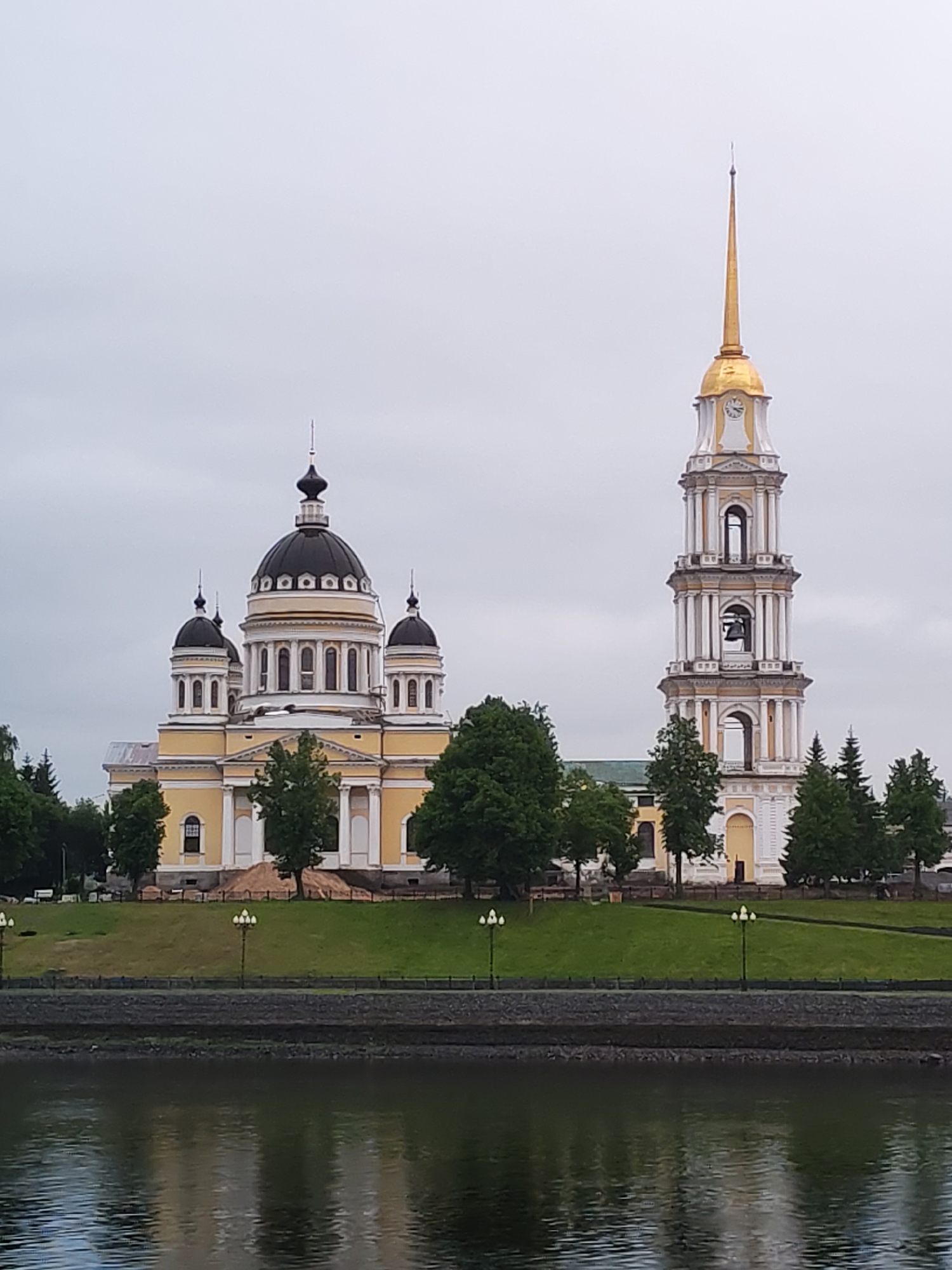 The image shows the Holy Trinity Cathedral in Bol'shie Mkhi, Russia, from across a body of water. The cathedral, a yellow and white building with dark domes, is located on a green embankment dotted with trees and lampposts. To the right of the cathedral is a tall, white bell tower with a gold dome and spire. The water reflects the buildings and sky. The sky is overcast.