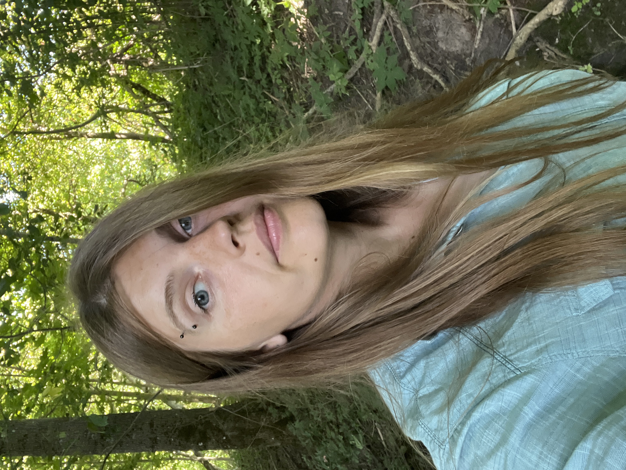 A young woman with long, light brown hair and blue eyes is photographed outdoors in a wooded area. She has a piercing above her left eyebrow. She is wearing a light blue, button-up shirt. The background is filled with trees and green foliage, suggesting a natural, possibly forested environment. The lighting indicates it is daytime, with sunlight filtering through the leaves.