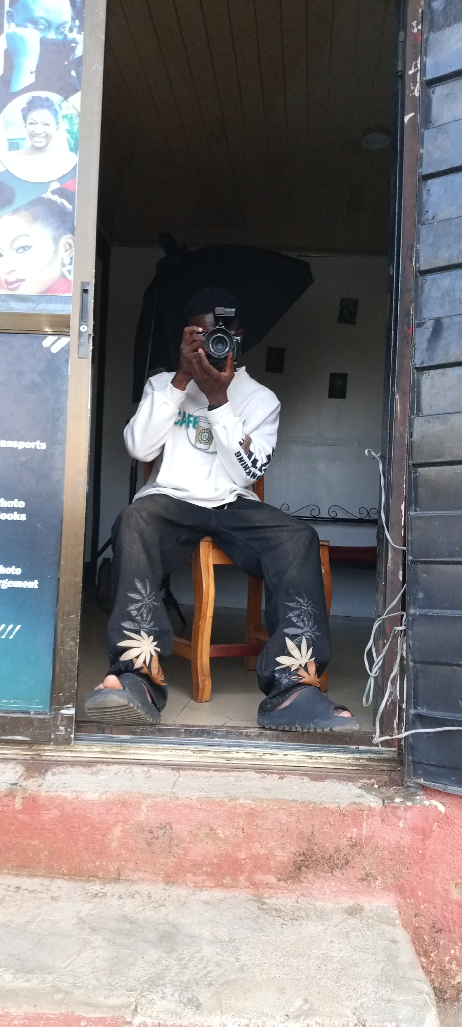 The image captures a young male photographer sitting on a wooden chair at the entrance of what appears to be a photo studio in Jos, Nigeria. He is holding a professional camera equipped with an external flash, posing as if about to take a picture, with the camera obscuring most of his face.
The photographer is wearing a white long-sleeved sweatshirt with a "CAFE" logo and a coffee cup graphic on the front, and text printed vertically down his right sleeve. He has black trousers adorned with prominent light brown/beige and grey leaf-like patterns on the lower legs, paired with dark slides or sandals. Behind him, inside the studio, a dark photography umbrella or softbox is visible, along with a light-colored wall and some framed pictures.
To the left of the entrance, a dark blue poster or banner advertises various photography services. Visible text includes "passports", "photo", "books", and "enlargement", accompanied by sample headshot photos of different individuals. The right side of the entrance features a dark metal door or frame with some white wires running vertically.
The ground immediately outside the studio consists of worn concrete steps, with sections painted or stained in reddish-brown, contrasting with patches of grey concrete. The overall setting suggests a small, local commercial establishment.