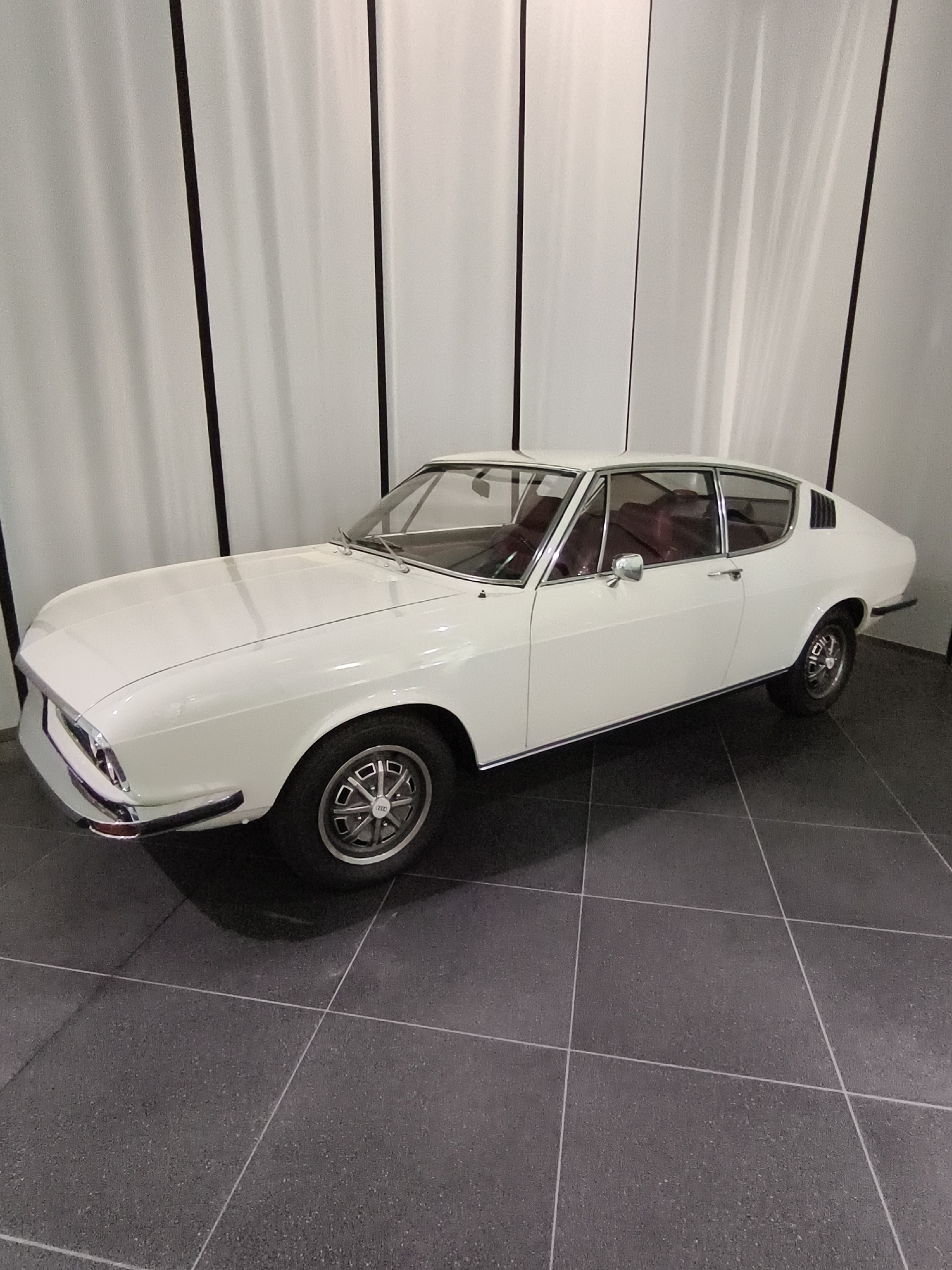 The image shows a white Audi 50 coupe on a black tiled floor in what appears to be a museum or exhibition space. The car is positioned at an angle, with its front left side closer to the viewer. The background is a white curtain with vertical black stripes, creating a sense of depth and highlighting the car. The car's interior, with its red seats, is partially visible. The Audi Forum Ingolstadt is a museum and event location that showcases the history of the Audi brand. This image was likely taken at the Audi Forum Ingolstadt, given the presence of an Audi vehicle and the likely setting of a museum.