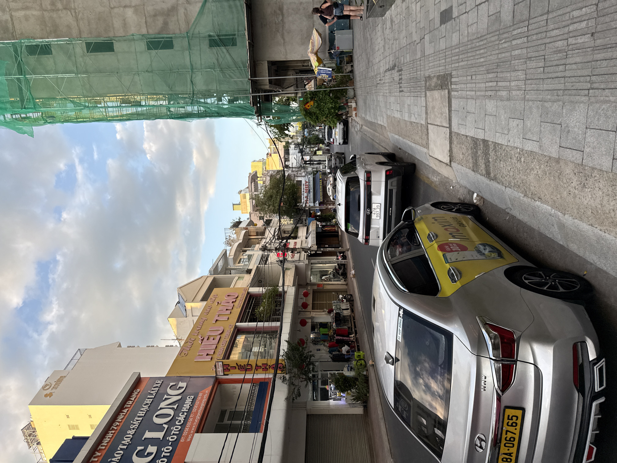 The image shows a street scene in Nha Trang, Vietnam. A silver sedan, identified as a Hyundai with license plate 84A-067.65, is parked on the right side of the street. It has a yellow decal with the Maxim logo and text "10%" on its rear passenger door. Two white SUVs are visible further down the street, one of which has a license plate ending in 97.39. The buildings lining the street are a mix of commercial establishments with brightly colored signs, including "LONG" which advertises driving lessons. On the right side of the street, a building is under construction, covered with green netting. Two individuals are standing on the sidewalk near the construction site, with one person appearing to be a woman wearing shorts and a tank top. The sidewalk is paved with distinct sections of large tiles and textured tactile paving. The sky is partly cloudy, and the overall lighting suggests it is late afternoon or early evening.