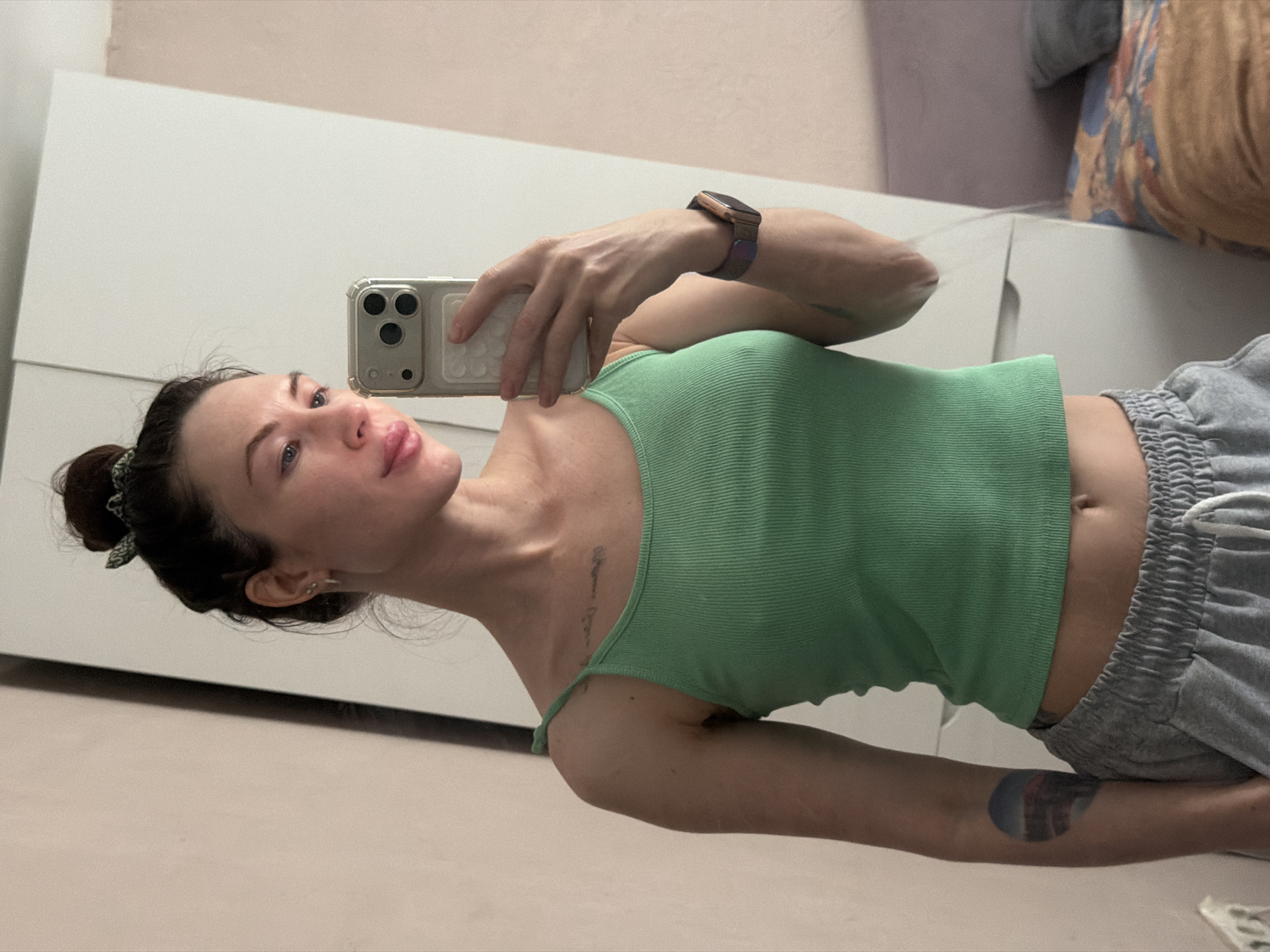 A woman is taking a selfie. She has brown hair tied in a bun, blue eyes, and fair skin. She is wearing a green ribbed tank top and grey sweatpants. She is holding a white smartphone in her right hand. She has a watch on her left wrist. There are tattoos on her left forearm and near her left collarbone. The background includes a white dresser, a lilac-colored wall, and a pillow with a pattern. There are no elements to suggest the location is Perm, Russia.