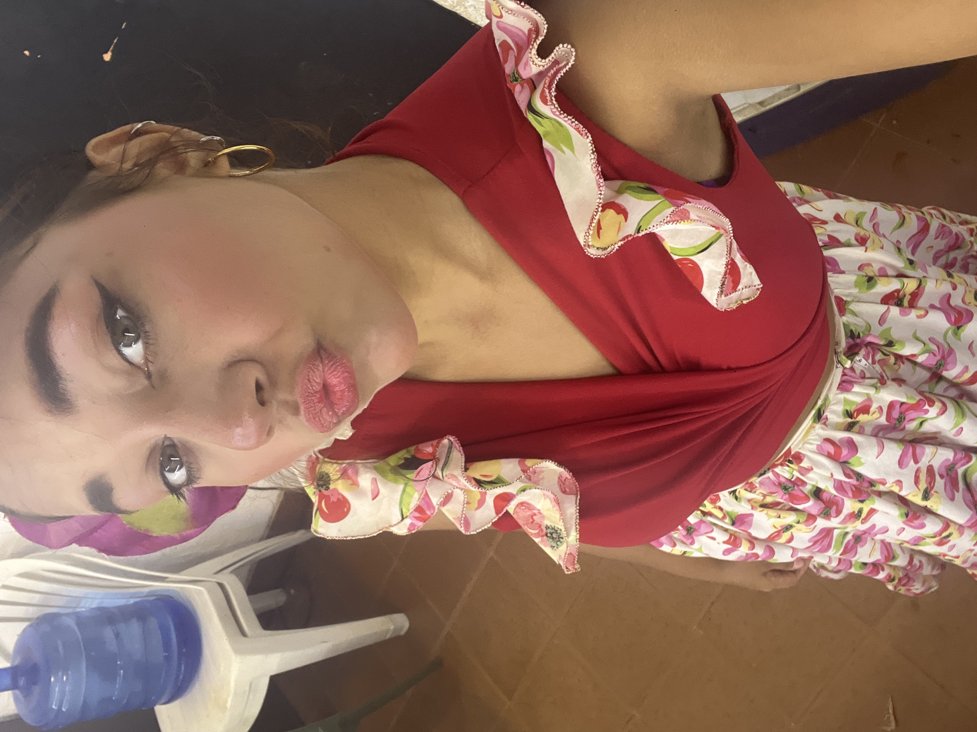 A young woman is captured in a selfie-style shot, posing with puckered lips in a "duck face" expression while looking up towards the camera. She has distinct eye makeup, including winged eyeliner, and prominent eyebrows. A gold hoop earring and several cartilage piercings are visible on her right ear. She is wearing a bright red sleeveless top featuring ruffled trim made from a white fabric patterned with colorful pink, green, yellow, and red flowers or fruit. Below the top, a white skirt or dress with a matching floral print is visible. The setting appears to be indoors, with a light brown tiled floor. To her left, a blue water bottle sits on a white plastic chair, and another white chair is partially visible to the right. A dark surface is behind her head. The image conveys a playful and casual atmosphere, situated in Porlamar, Venezuela. No visible text is present.
