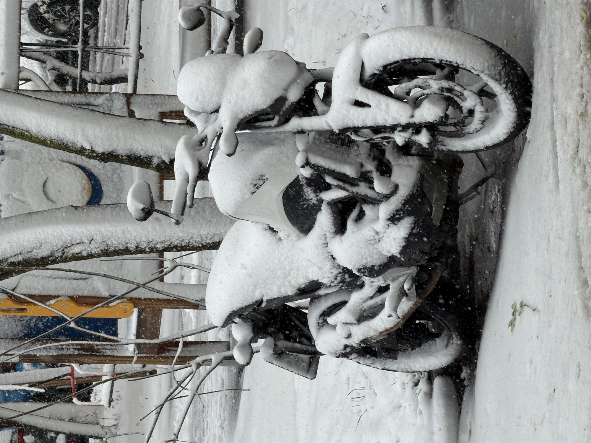 A motorcycle is heavily covered in snow. It appears to be parked in a residential area in Khimki, Russia, given the presence of what seems to be a children's playground structure visible in the background to the left. The ground, surrounding structures, and the motorcycle itself are blanketed in a thick layer of fresh snow.