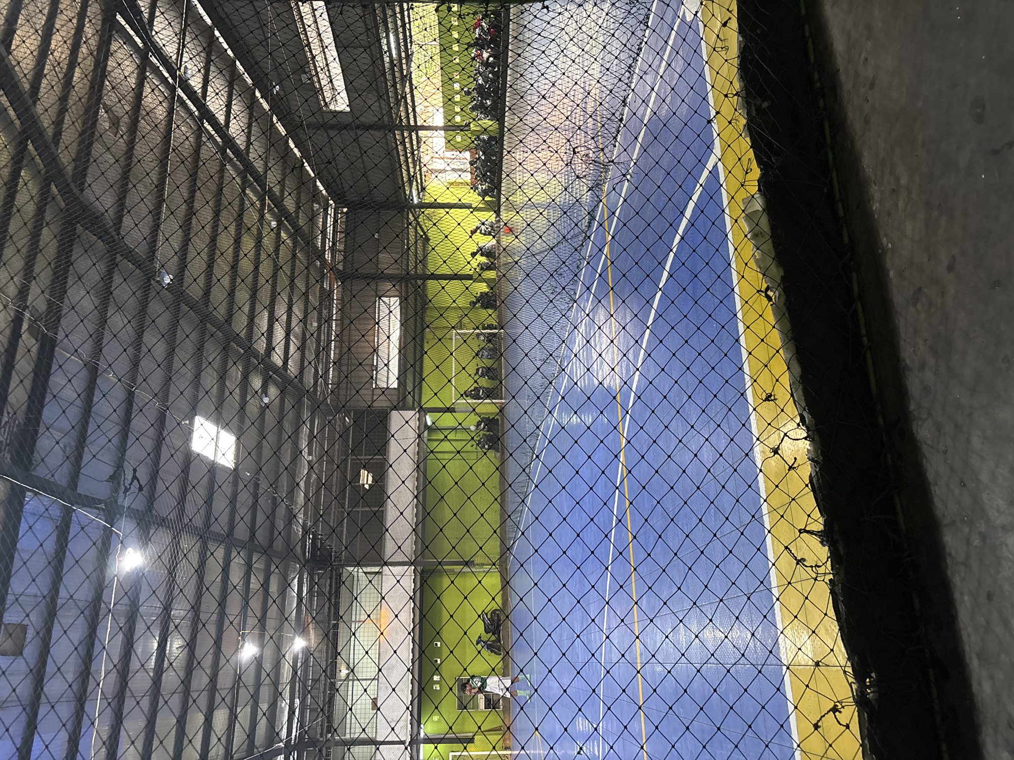 A futsal court is depicted through a black net, inside a covered facility. The court is blue with yellow and white markings. The walls of the facility are lime green, and a row of motorcycles is parked along the far wall. A person in athletic wear is visible on the court. The ceiling of the facility is covered with corrugated metal and features lighting fixtures. The overall environment suggests an indoor sports complex. This image was taken in East Jakarta, Indonesia.