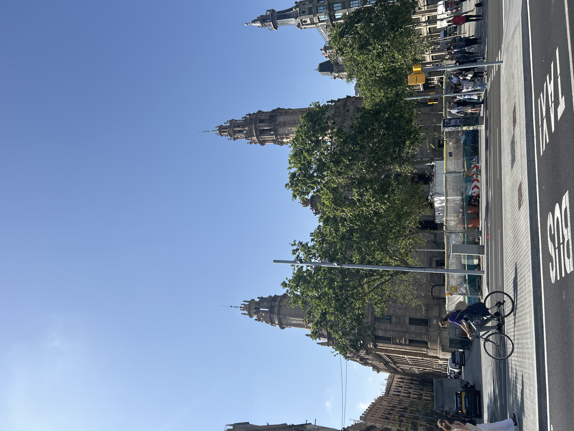 The image depicts a bright, sunny urban scene in Barcelona, featuring grand classical architecture, lush trees, and a busy street. The sky is a clear, vibrant blue with no visible clouds.
On the left side of the frame, several magnificent, ornate buildings rise, characterized by their detailed facades, multiple stories, and decorative towers or spires. These structures are partially enveloped by the dense, green canopies of numerous mature trees, suggesting a tree-lined avenue or park adjacent to the buildings.
The right side of the image features a wide paved street with clear white road markings. The word "TAXI" is prominently painted in large letters on the asphalt, and a partial view of "BUS" can also be seen, indicating designated lanes. A person wearing a purple top and dark pants is actively cycling on a dark-colored bicycle along this street, heading towards the left. Further down the road, several vehicles, including cars and a yellow taxi, are present.
Pedestrians are visible on the sidewalks and crosswalks. In the midground, near the base of the ornate buildings, groups of people are walking and standing. Street furniture, such as lampposts and a yellow traffic light signal box, are interspersed throughout the scene. Orange and white construction barriers are also visible in front of one of the buildings, indicating ongoing urban development or maintenance. The overall impression is one of a lively city environment bathed in clear daylight.