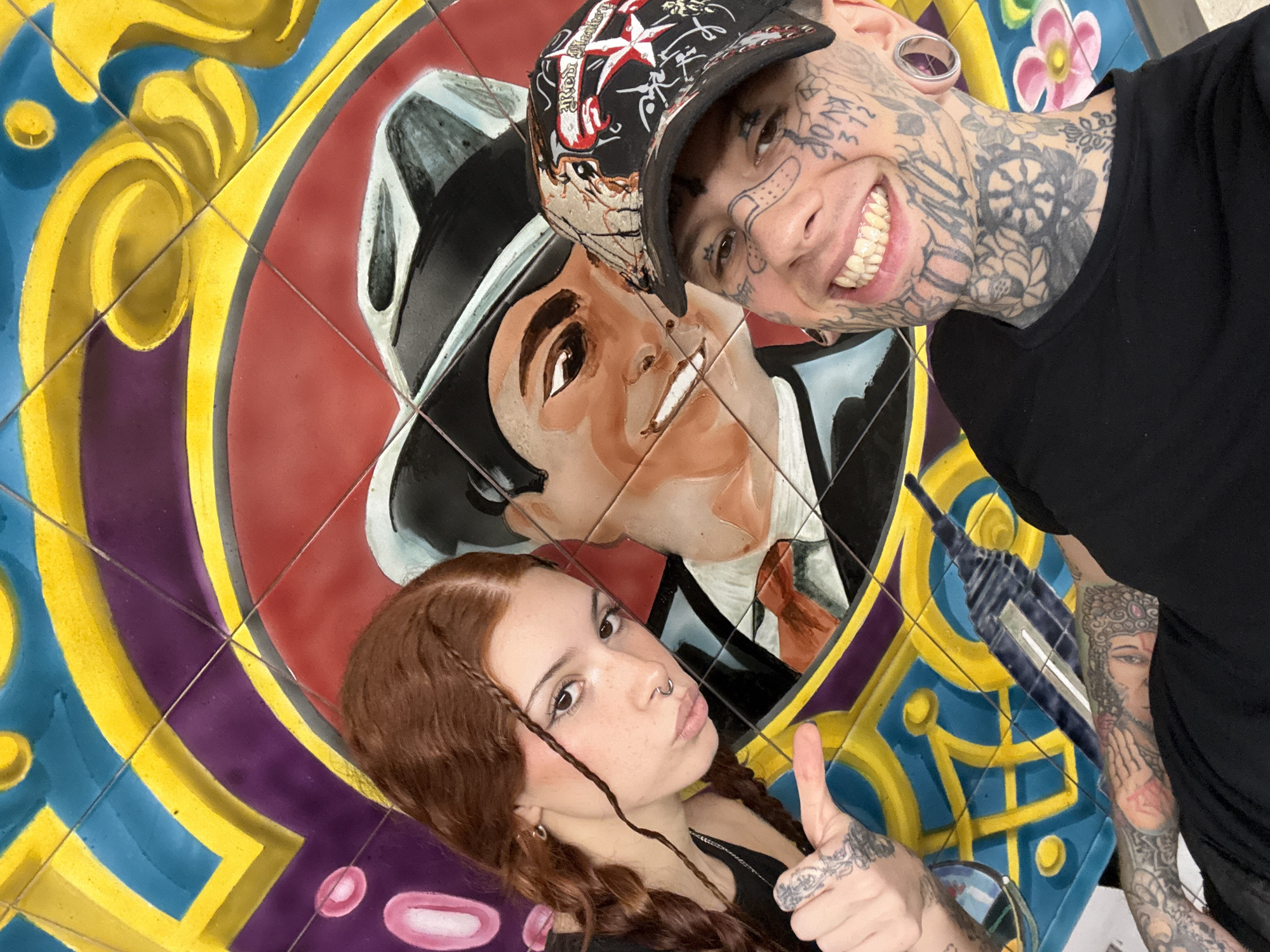 The image depicts two people posing in front of a tiled mural. The mural appears to be a stylized portrait of a man in a suit and fedora, surrounded by colorful geometric patterns. The woman on the left has auburn hair, and she is giving a thumbs-up. The man on the right has extensive facial and neck tattoos and is wearing a baseball cap.
Based on visual clues (the artistic style of the mural, the attire, and architecture), the location may be in the city of Buenos Aires, Argentina.