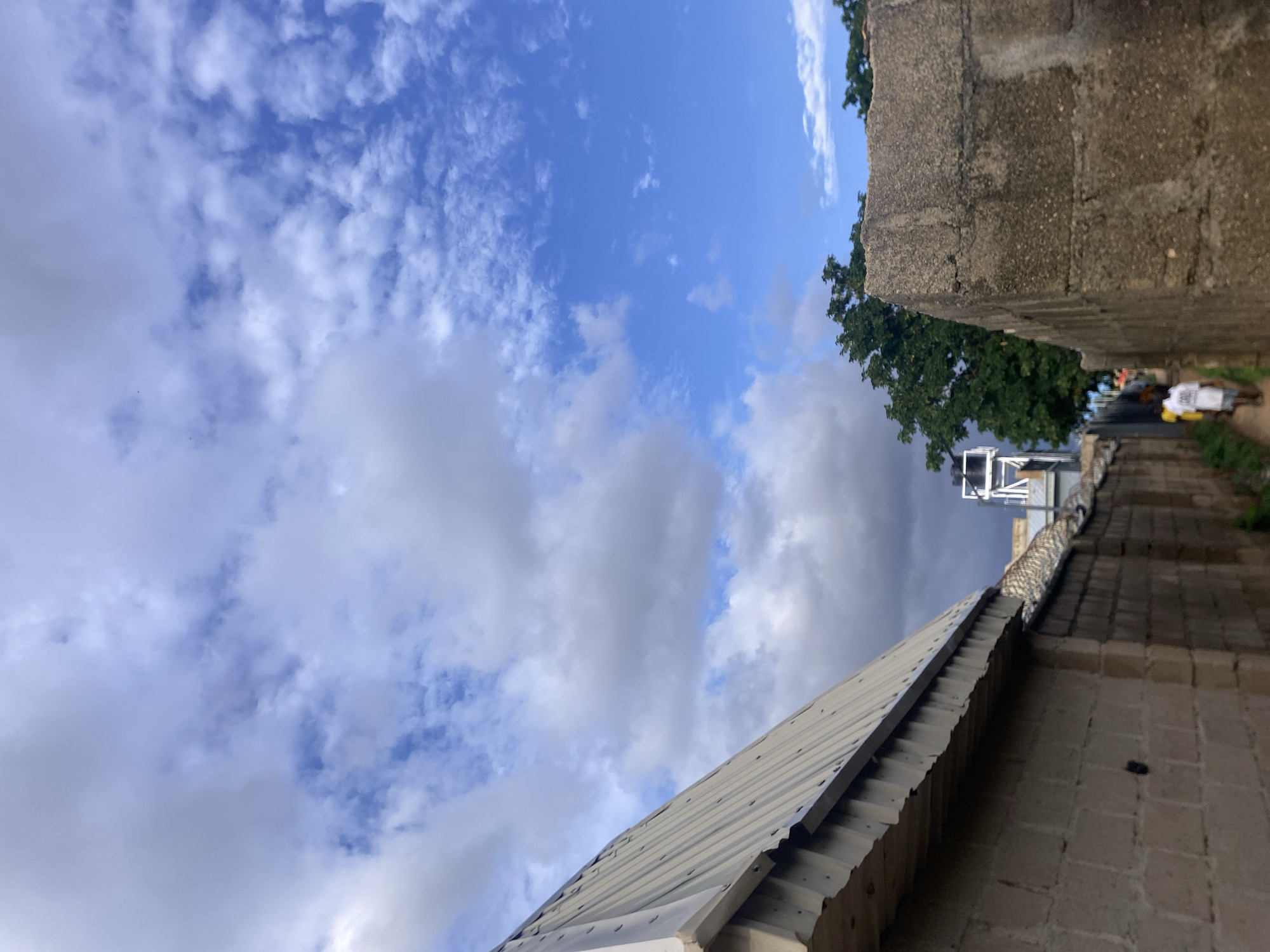 The image depicts a narrow alleyway or passage within the city of Jalingo, Nigeria, under a sky with a mix of blue patches and scattered white and grey cumulus clouds. On the left, a light-colored corrugated metal roof slopes downwards over a light-toned concrete block wall. Below the roof's edge, deep shadow is present. Further along this left wall, a security fence, possibly featuring barbed or razor wire, is visible atop a lower structure. The right side of the passage is defined by a tall, rough-textured, dark grey concrete block wall. In the mid-ground, down the alleyway, several individuals are visible walking away from the viewer, with one person in a white t-shirt and light trousers being most distinct. Beyond the structures and people, a prominent green tree stands against the sky. In the far distance on the left, a tall, white metal tower, potentially a water tower or similar utility structure, is visible. The scene is illuminated by natural daylight, creating visible shadows.